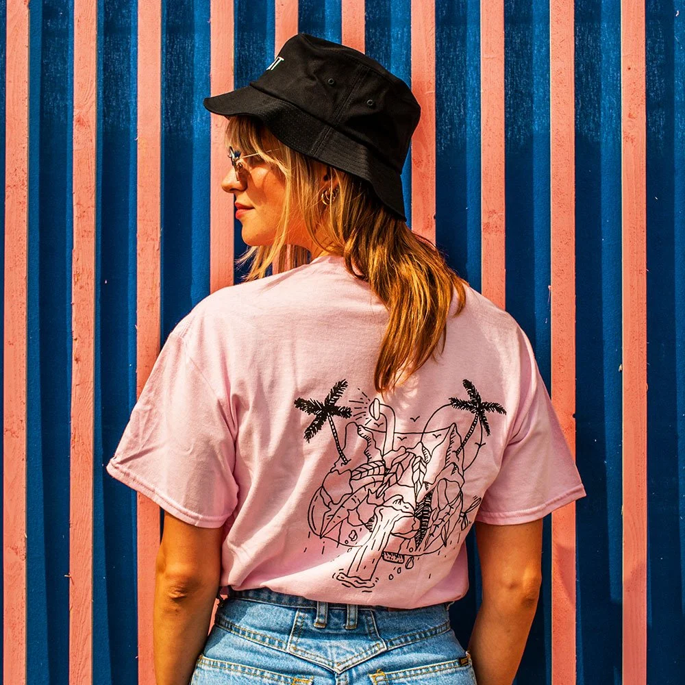 Een vrouw met een zwart hoekhoedje en een roze T-shirt met een grafische tekening van een hond en palmbomen, staand voor een achtergrond met blauwe en roze verticale strepen.
