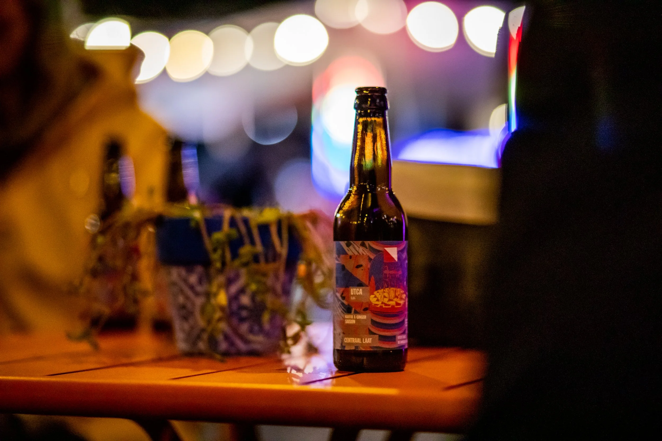 Een flesje Brouwerij Eleven bier op een houten tafel in de tuin van het Centraal Museum Utrecht op de Utca's Finest X Centraal Laat 'de ommuurde stad' avond, met kleurrijke, wazige achtergrondverlichting. Ontwerp bierlabel door Temple Isle Trademark