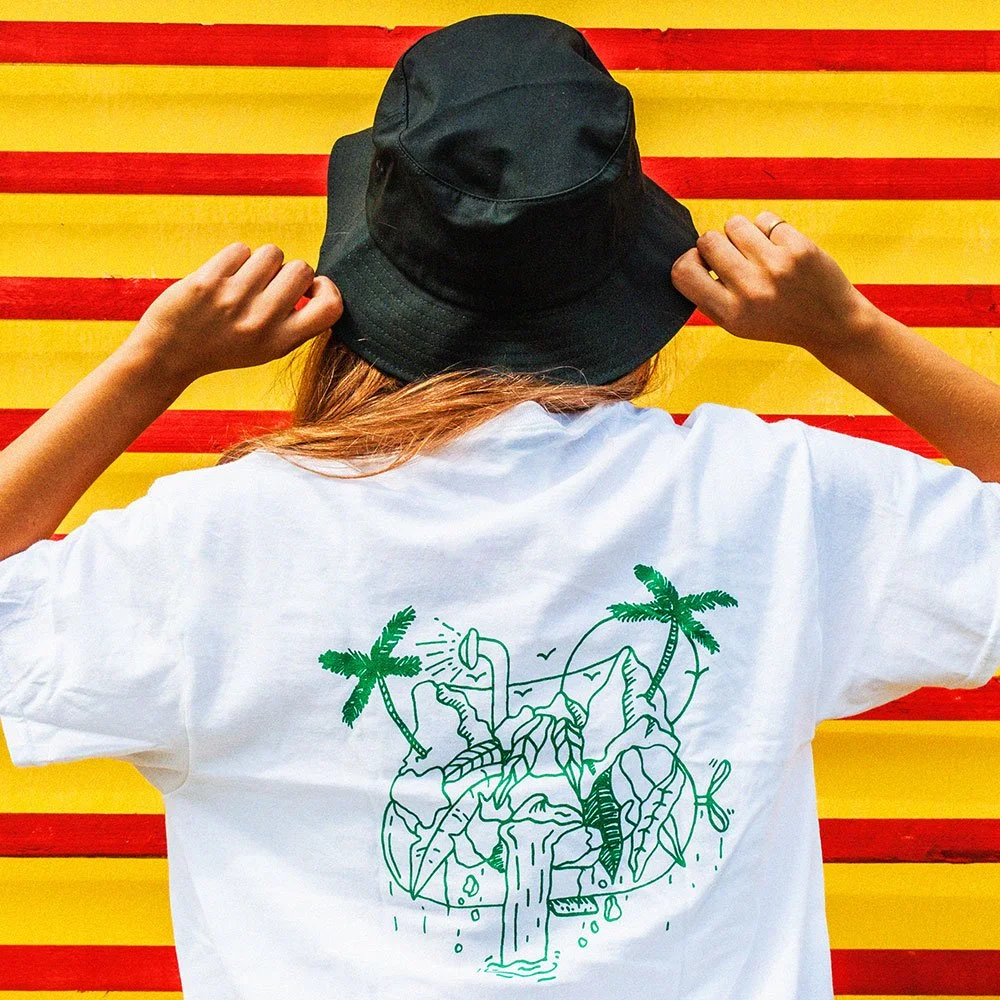 Person met witte T-shirt met groene illustratie van een jungle met palmbomen en een waterval, draagt een zwarte hoed, staat voor een muur met rode en gele strepen.