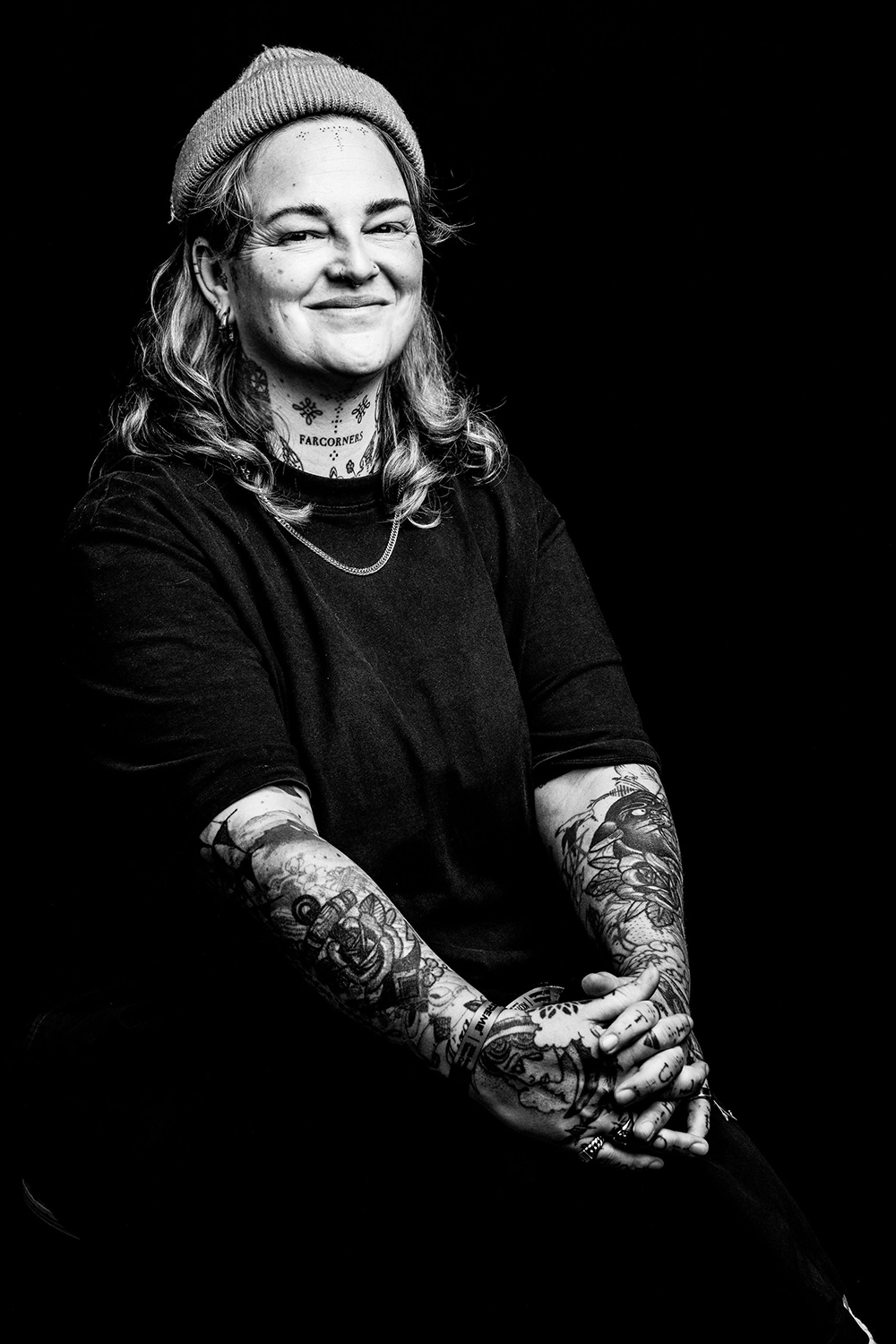 Zwart-wit foto van een vrouw met tattoos, een beanie en een korte mouwen shirt, lachend en zittend met gekruiste handen.
