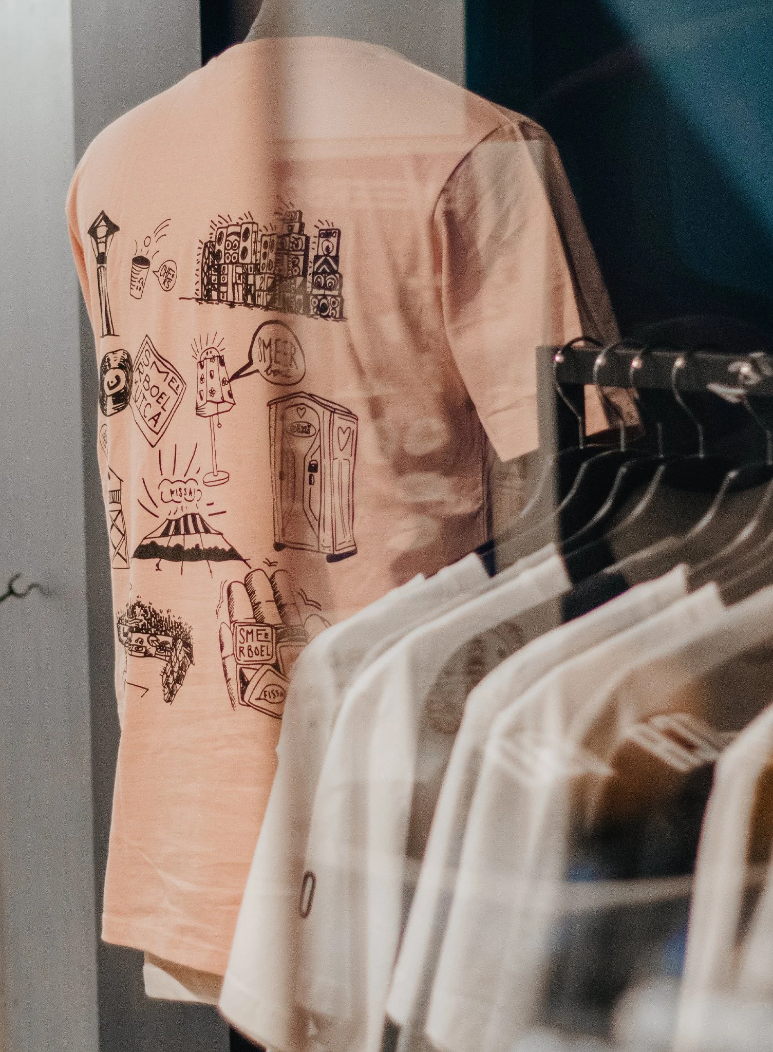 Close-up van een persoon die een roze T-shirt draagt met zwart-witte tekeningen en tekst, naast een witte kledinghanger met deels zichtbare witte kleding.