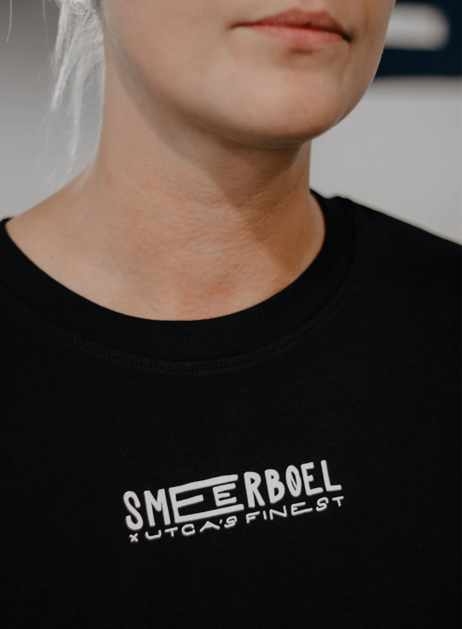 Close-up van een persoon die een zwarte T-shirt draagt met witte tekst. De tekst op het T-shirt leest 'SMEERBOEL' bovenaan, met andere woorden eronder.