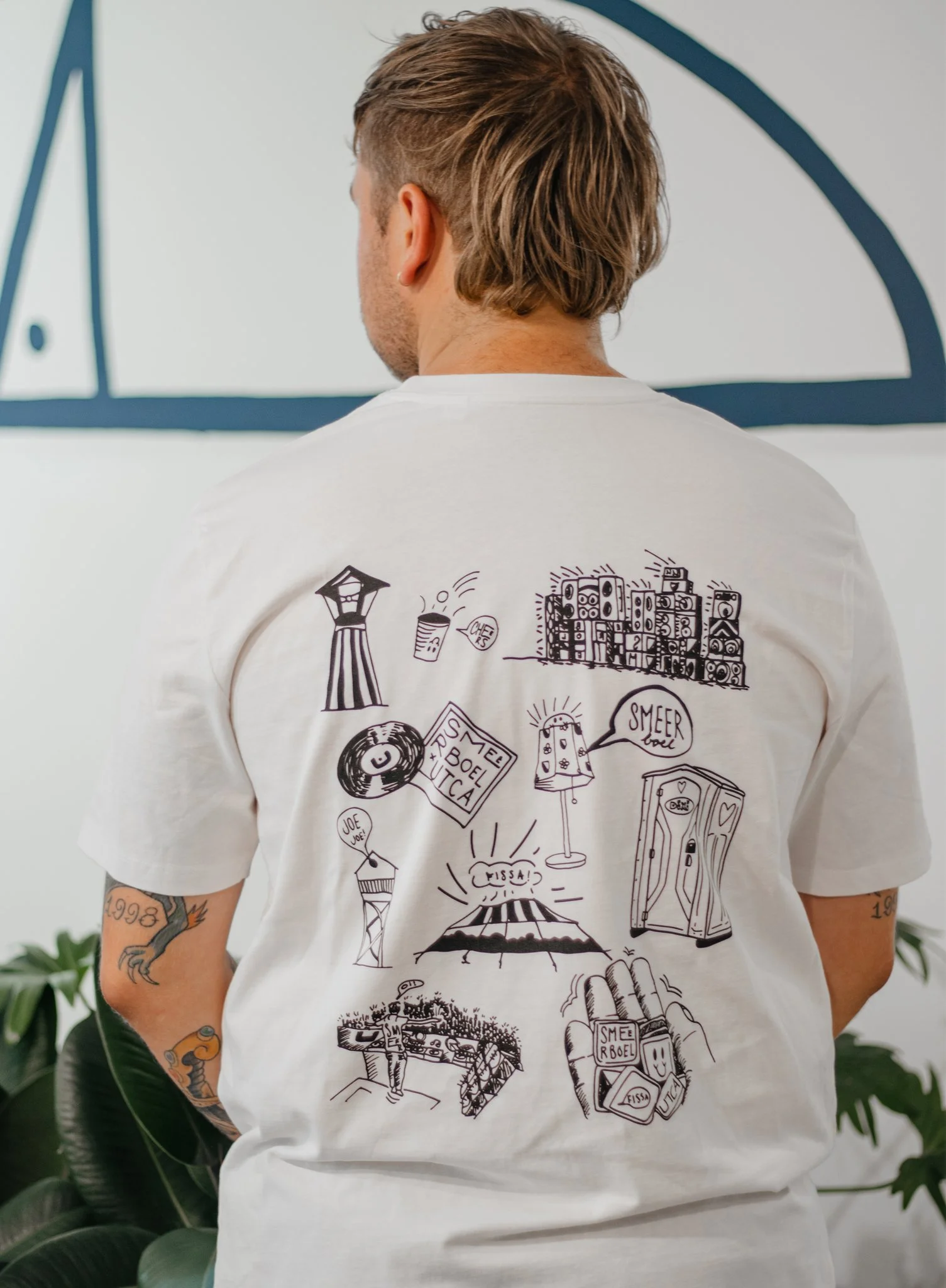 Man met tattoos op armen en blond haar, gezien van achter, draagt een witte t-shirt met zwart-witte illustraties en tekst, staat voor een witte muur met blauwe decoratie.