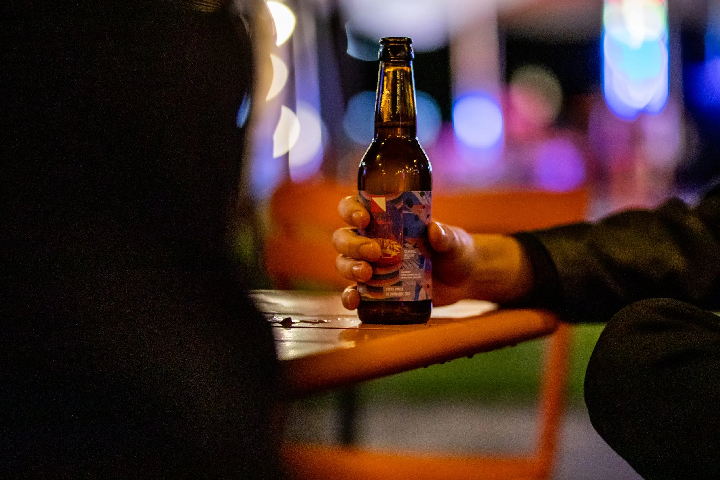 Persoon die een bierflesje vasthoudt in een donker verlichte omgeving met kleurrijke lichten op de achtergrond. Centraal Museum Utrecht