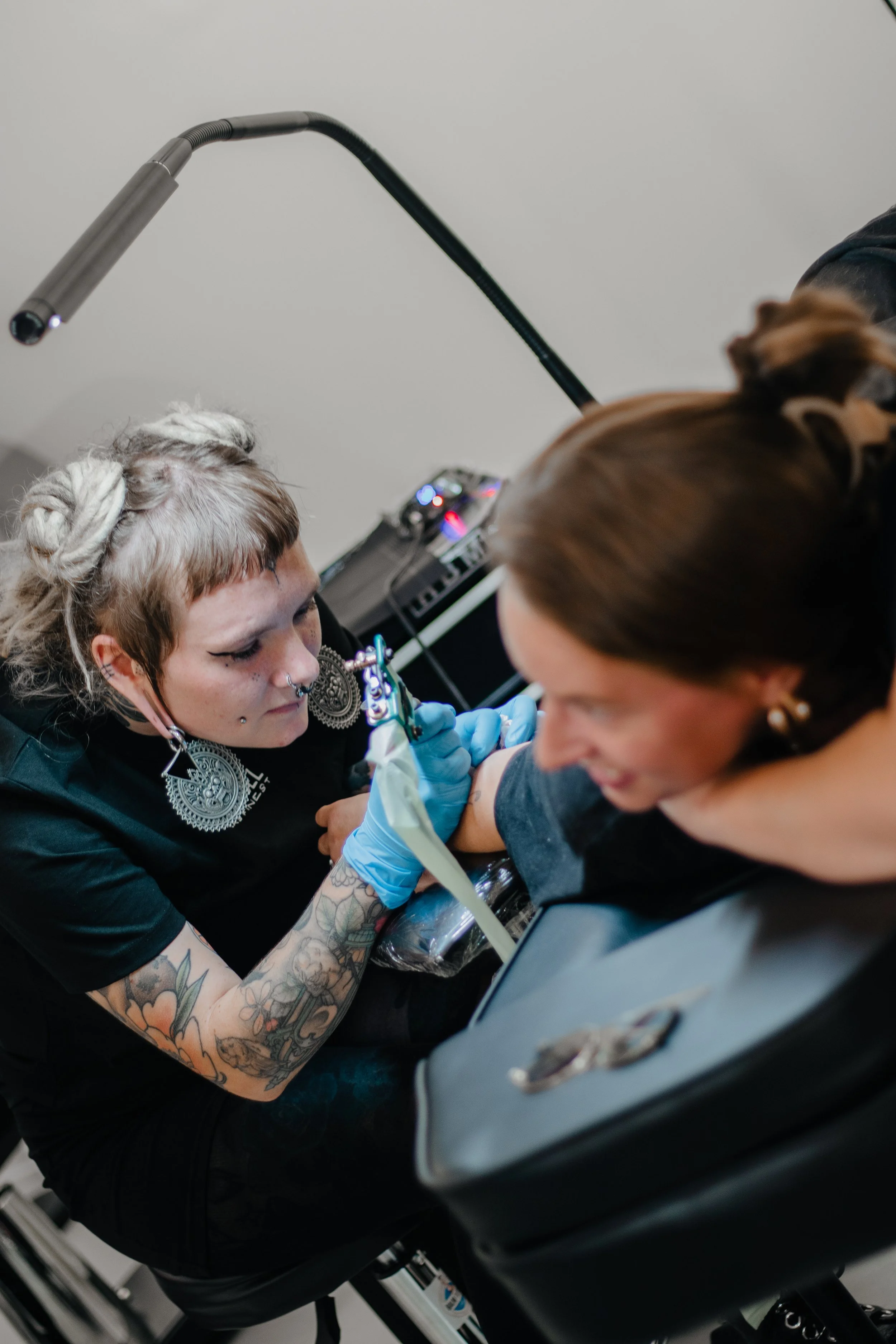 Tattoo- en piercingshouder geeft tatoeage aan een cliënt. De tatoeëerder heeft kort haar en grote oorbellen, en is bezig met het inkleuren van een tatoeage op de arm van de cliënt.