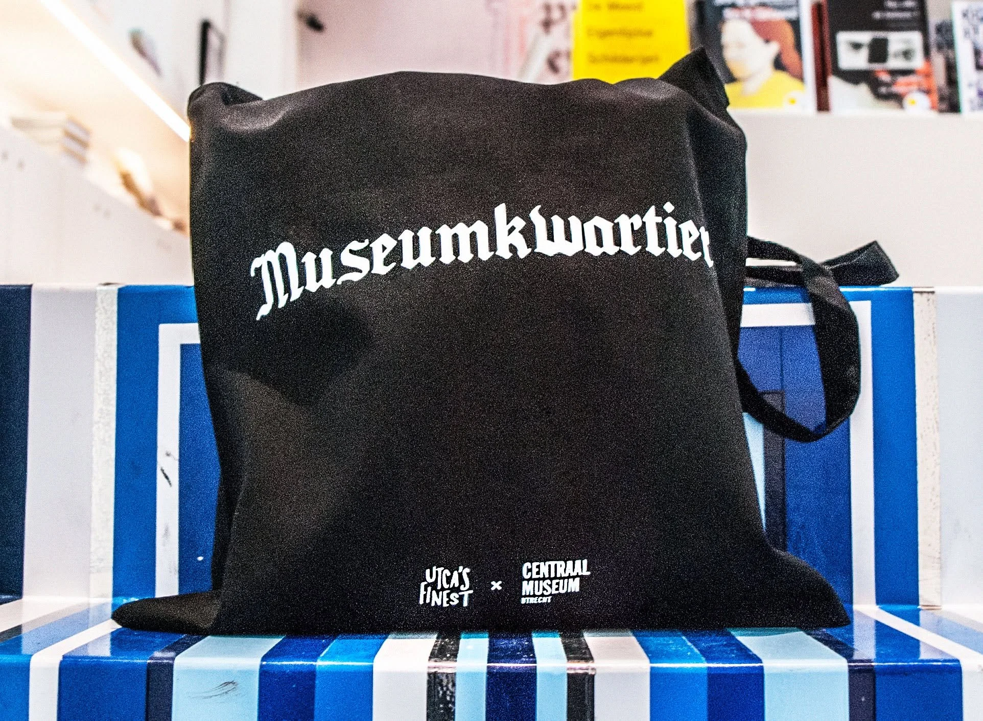 Zwarte tas met witte tekst 'Museumkwartier' en logo's van Lucas's Finest en Central Museum. De tas ligt op een blauwe en witte gestreepte tafel, met een brede achtergrond vol boeken en posters.