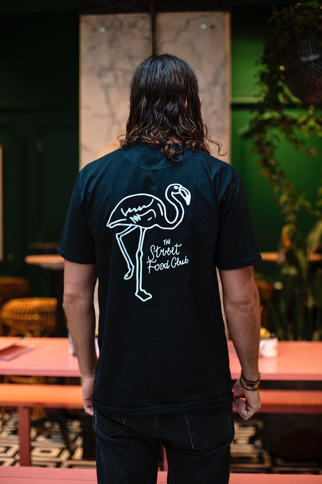 Man met lang, wavy haar dat naar de achterkant kijkt, draagt een zwart T-shirt met een witte flamingo-tekening en de tekst 'The Street Food Club' erop, binnen een restaurant of café.