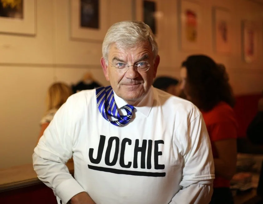Man met witte shirt met de tekst 'Jochie' en een blauwe strik, in een restaurant of café, met andere mensen op de achtergrond.