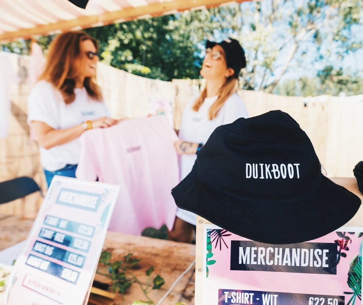 Een zwarte hoed met de tekst 'DUIKBOT' ligt op een tafel bij een merchandise kraam met een bord dat prijzen toont, op de achtergrond zien we twee vrouwen die samen kleding bekijken, en een bord met een prijslijst.