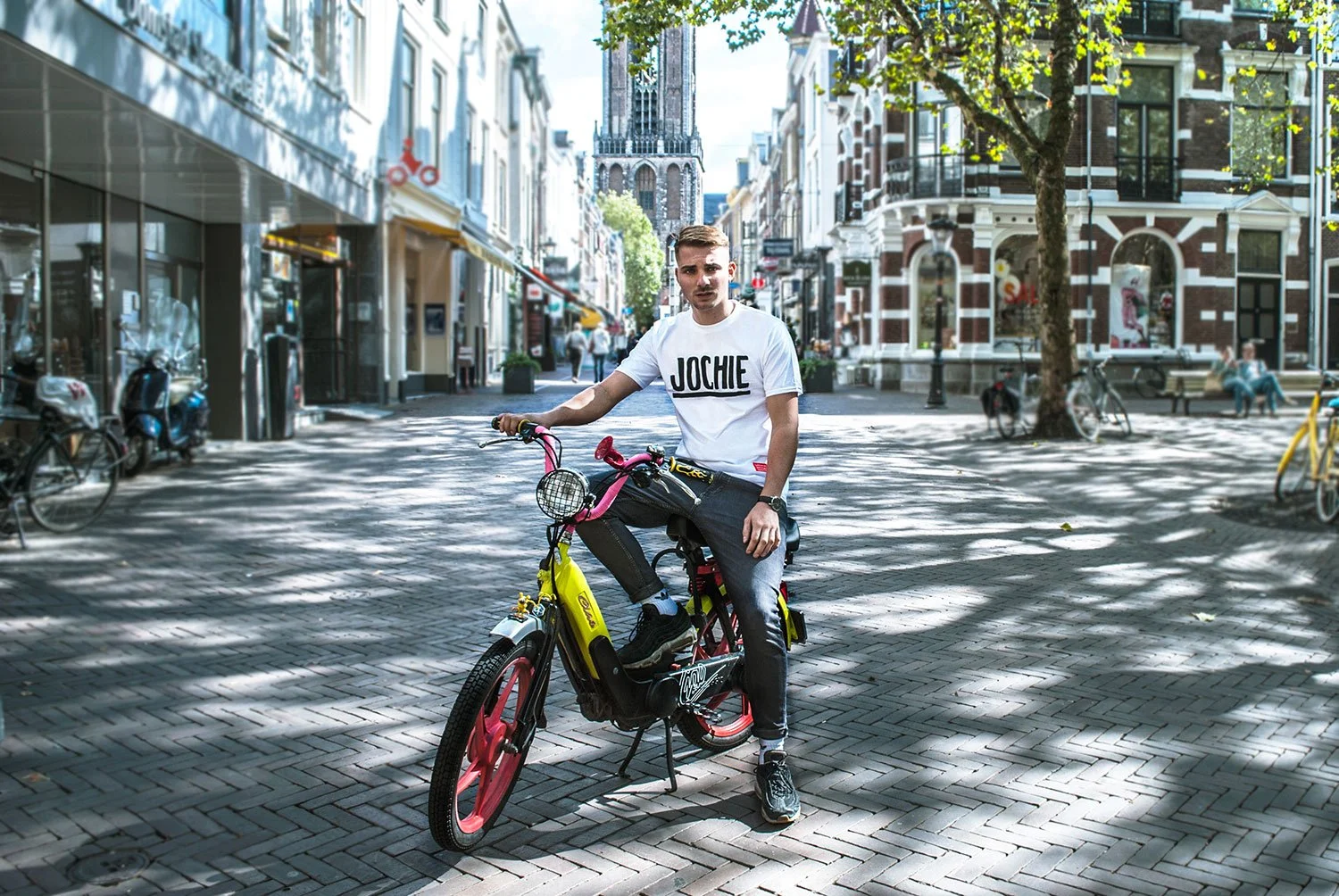 Jongeman op een fietsen in een stadscentrum met historische gebouwen en een toren op de achtergrond, schaduw van bomen en mensen op de achtergrond.