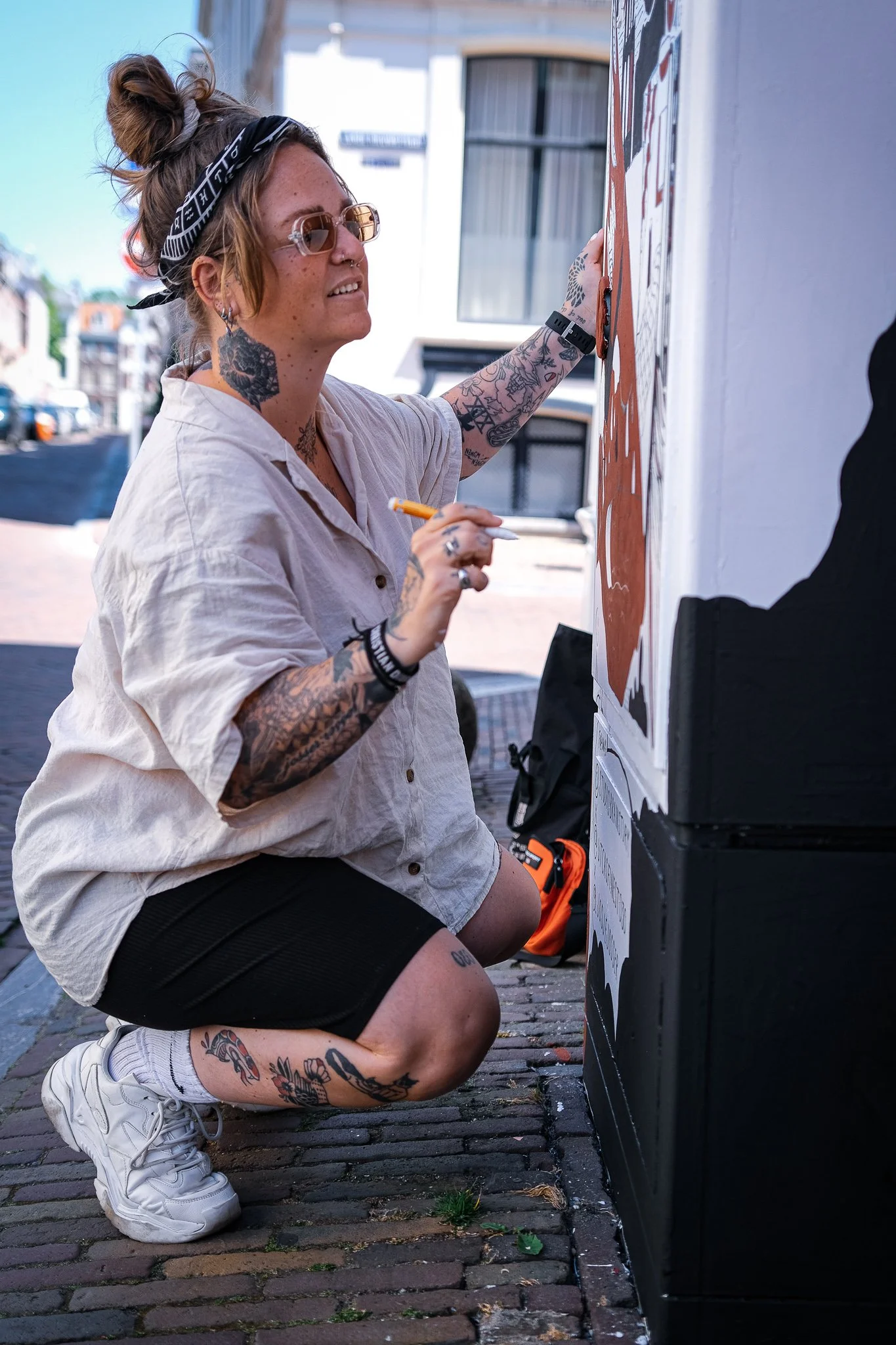 Een vrouw met tattoos en een haarband knielt neer terwijl ze iets op een muur schildert, buiten op een straat. Thirty030 vraagt mij of ik een ontwerp wil maken voor een electriciteitskastje, van kastje tot canvasje. Door Swaak, Gemeente Utrecht