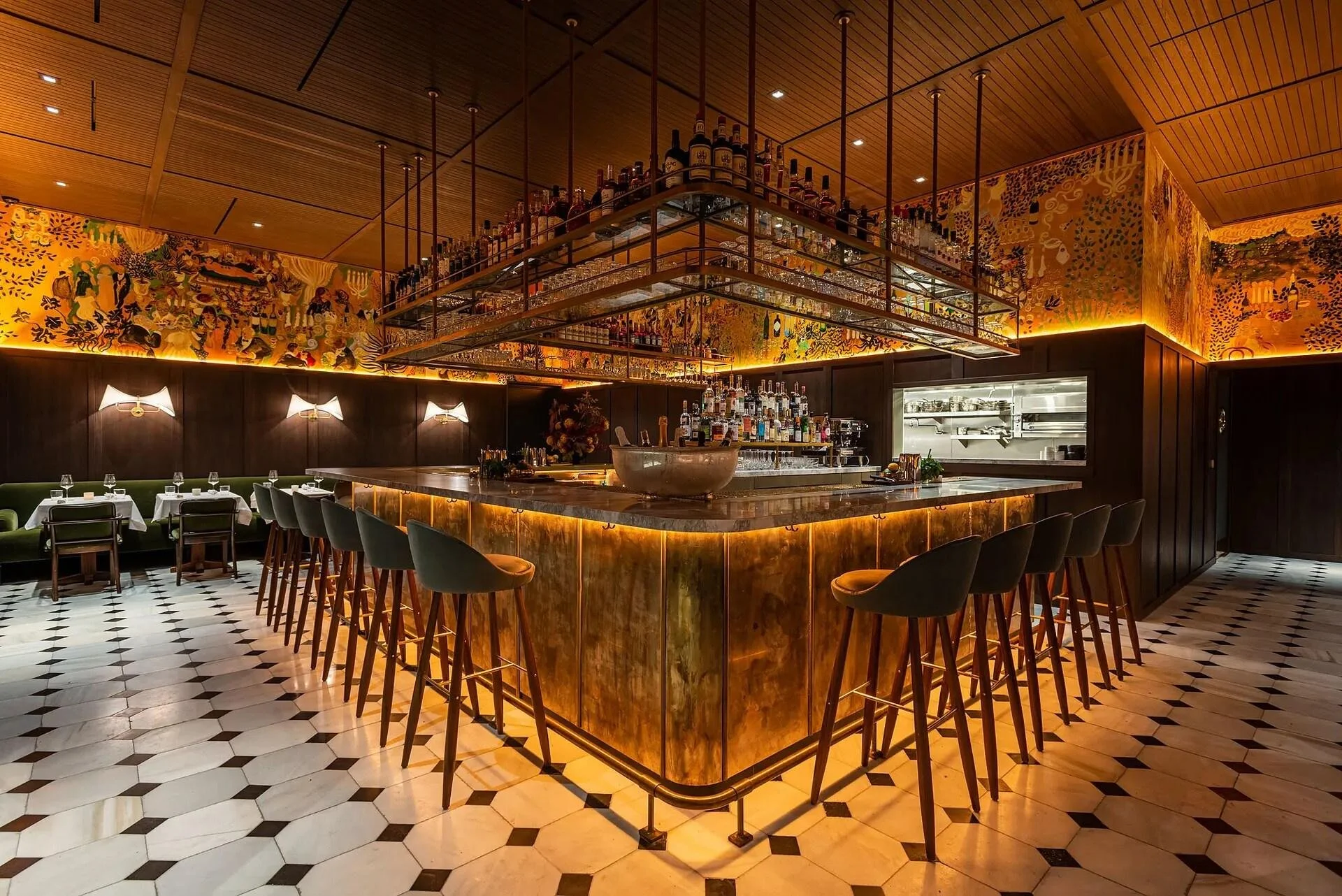 Interior of a modern bar with a marble-topped counter, bar stools, and a suspended shelf displaying various liquor bottles, with warm lighting and decorative mural walls.