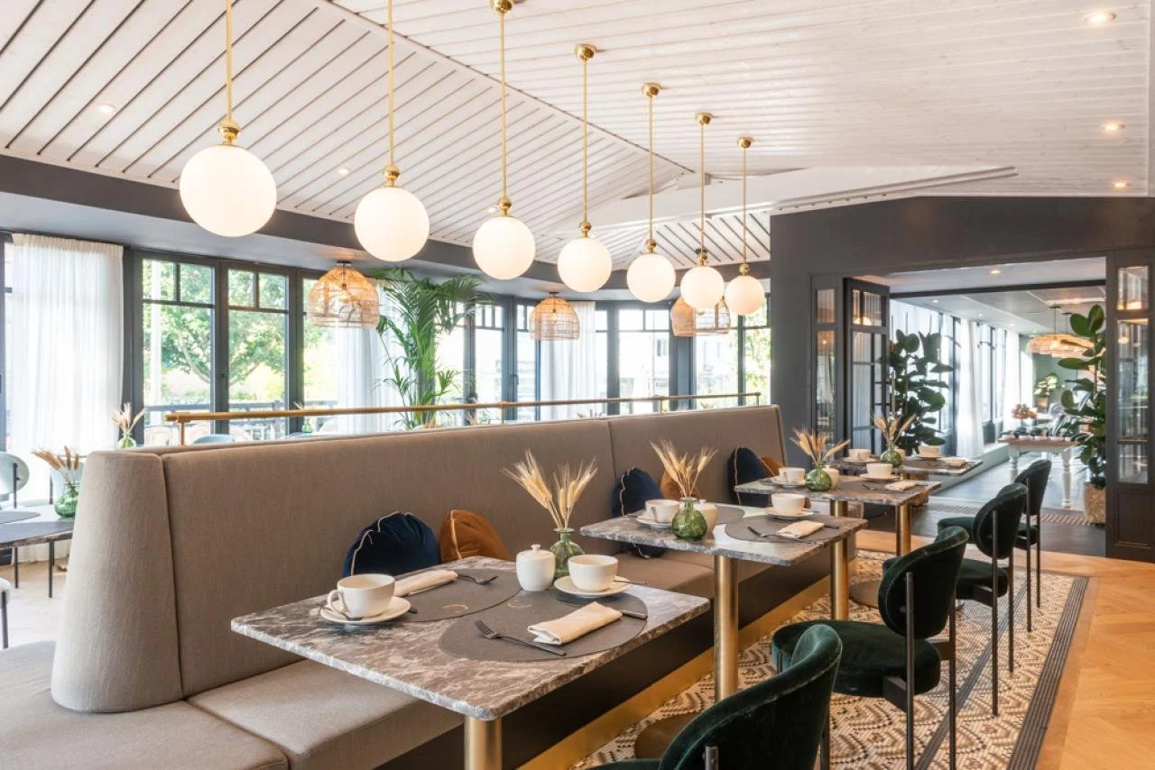 A modern restaurant interior with marble-top tables, velvet chairs, and large windows with curtains, decorated with green plants and table settings.
