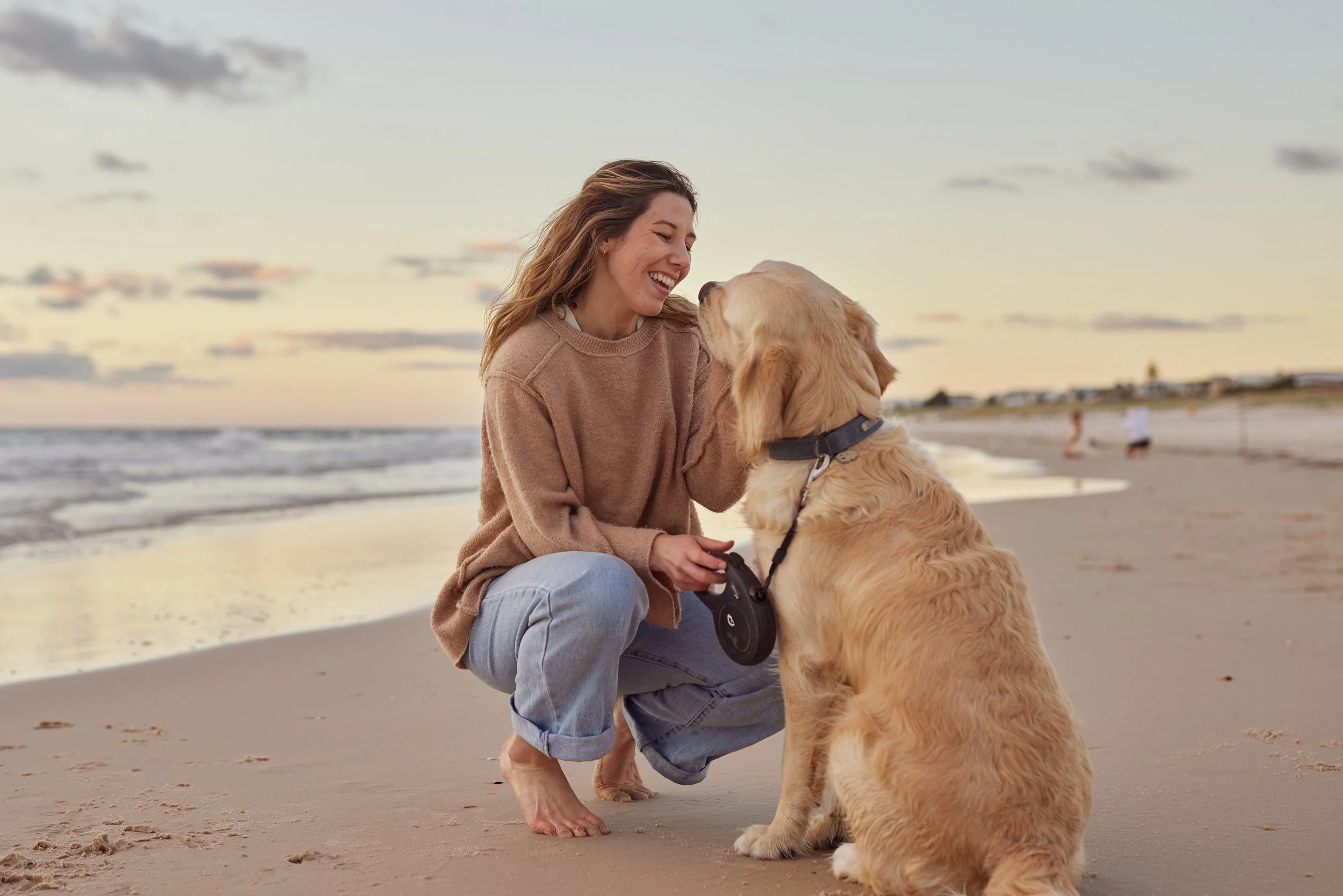 A woman with long hair, Darcie, wearing a beige sweater and jeans, smiling at a golden retriever on a beach.