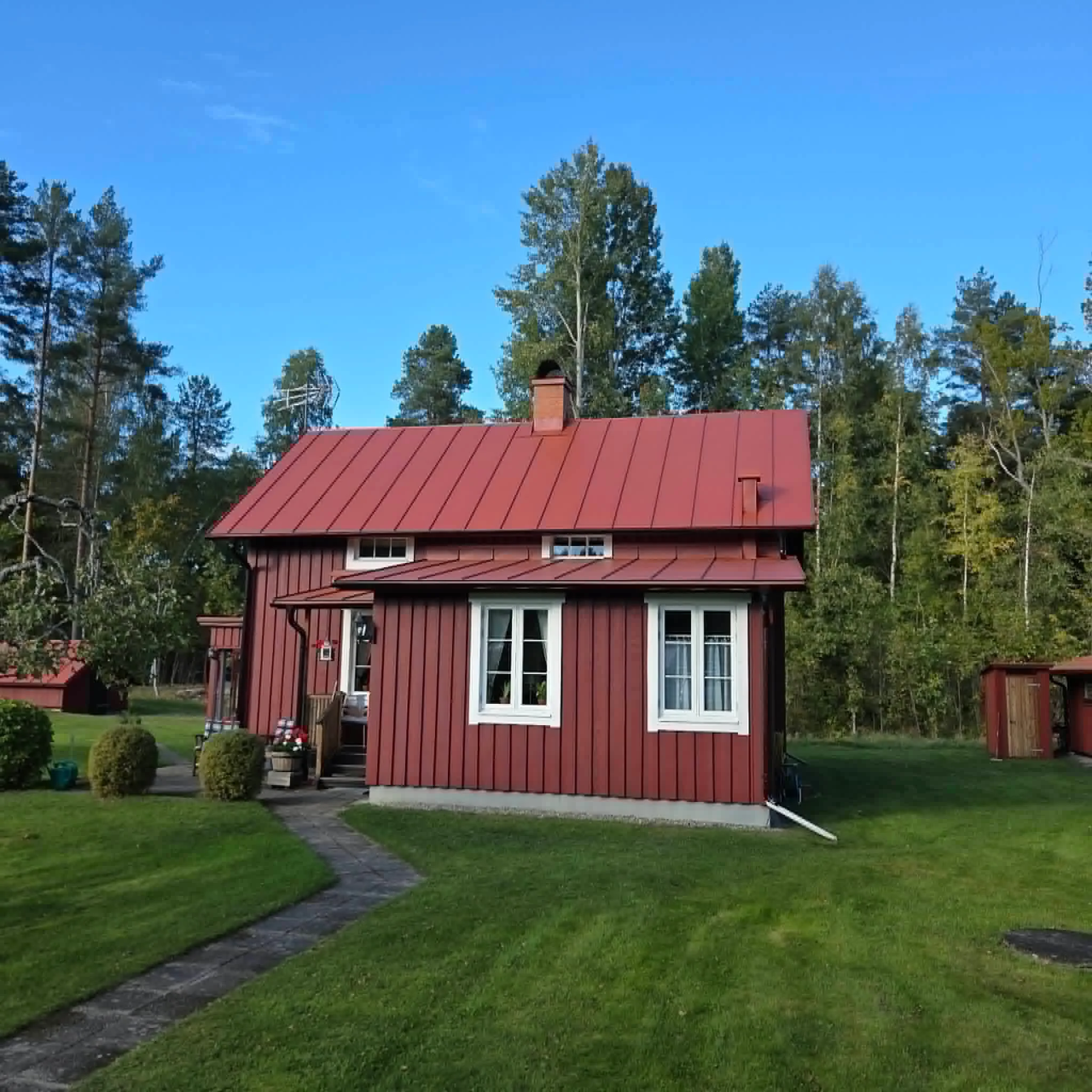 Hus med rött tak omgivet av grönt gräs och träd i bakgrunden under en klar blå himmel.