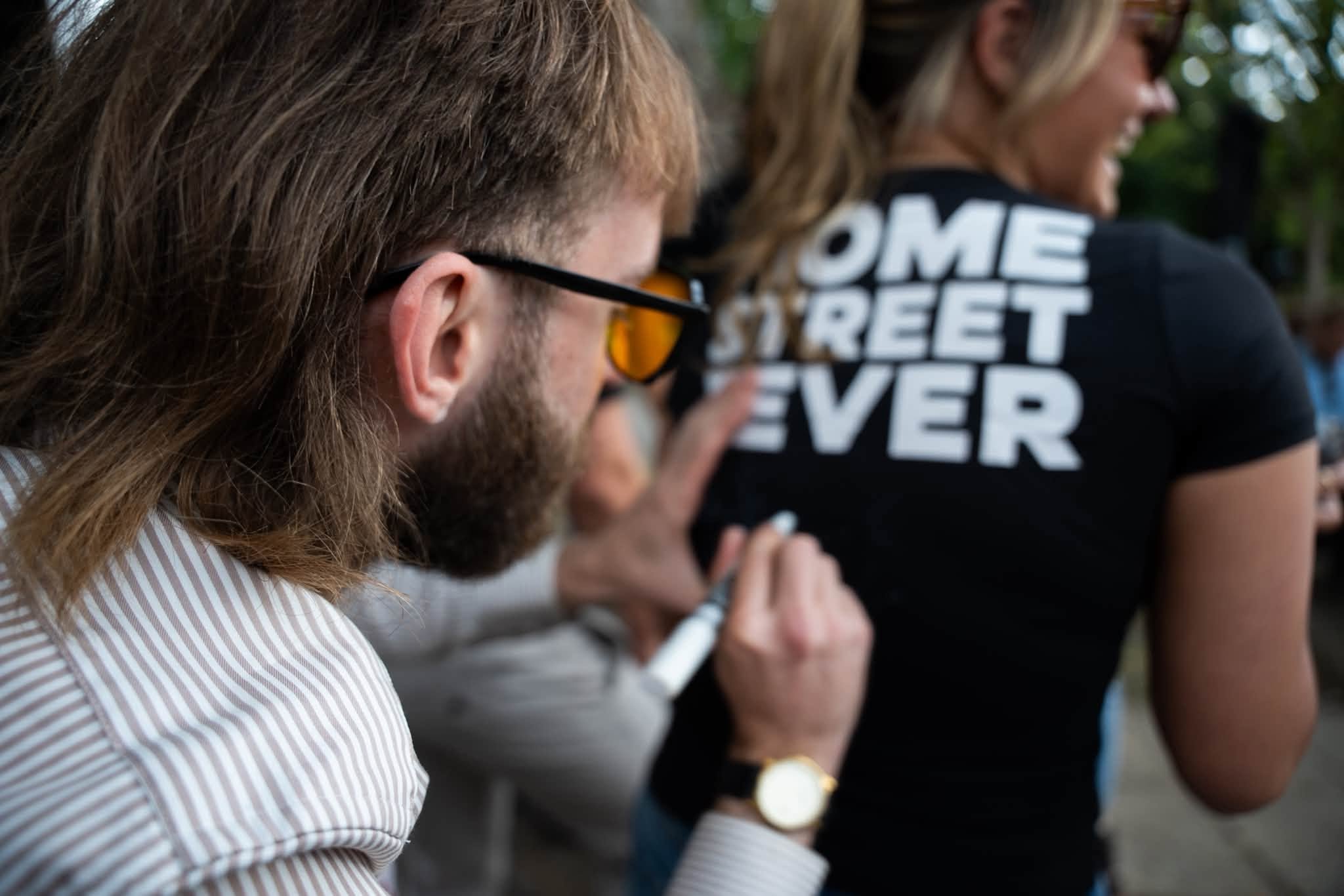 Close-up of two women attending a gathering outdoors, focusing on the woman with glasses and long brown hair in a striped shirt in the foreground, and a woman in a black shirt with white text in the background.