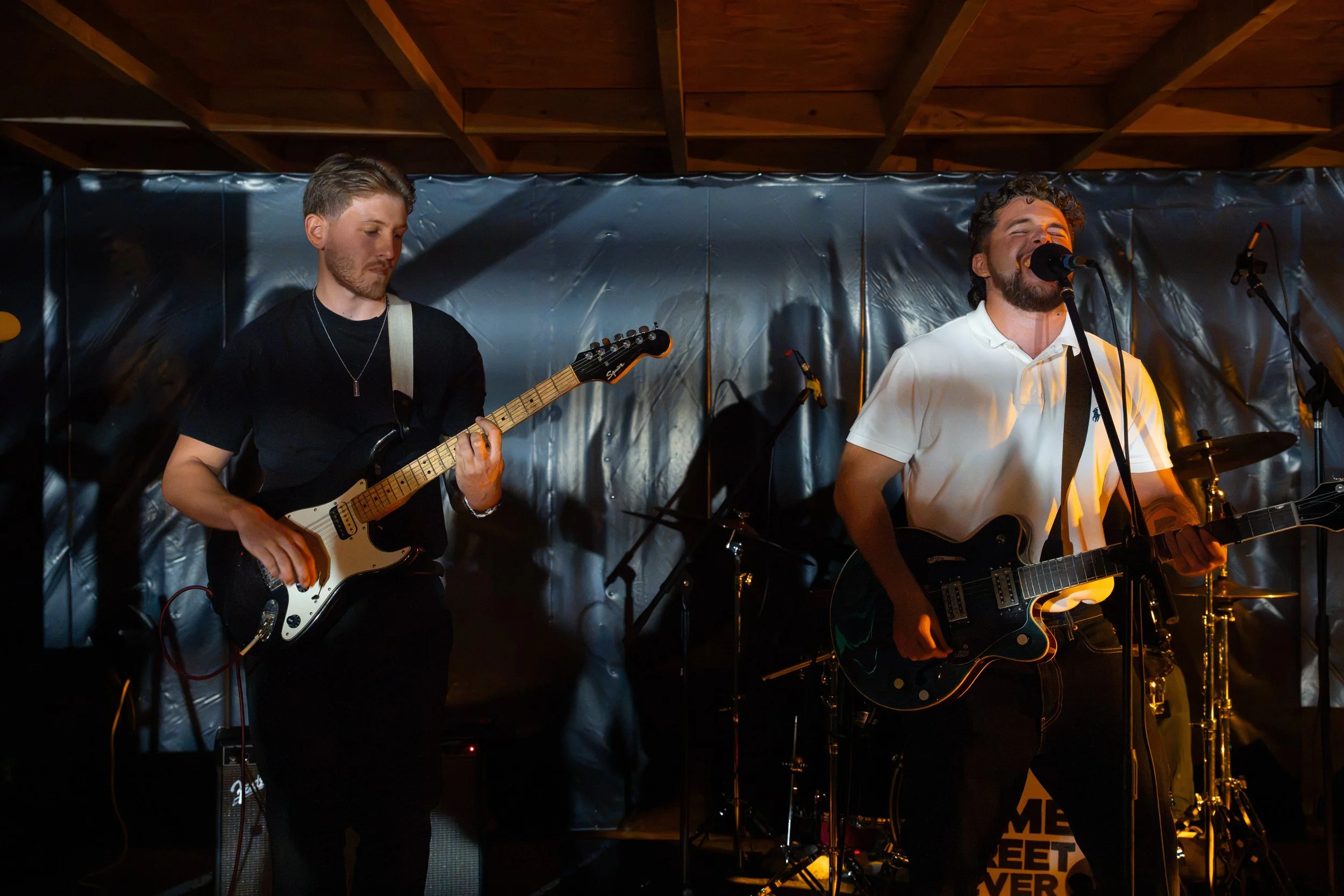 Two male musicians performing on stage; one playing an electric guitar with a black strap, the other singing into a microphone while playing an electric guitar, surrounded by musical equipment, with a dark backdrop and a wooden ceiling.