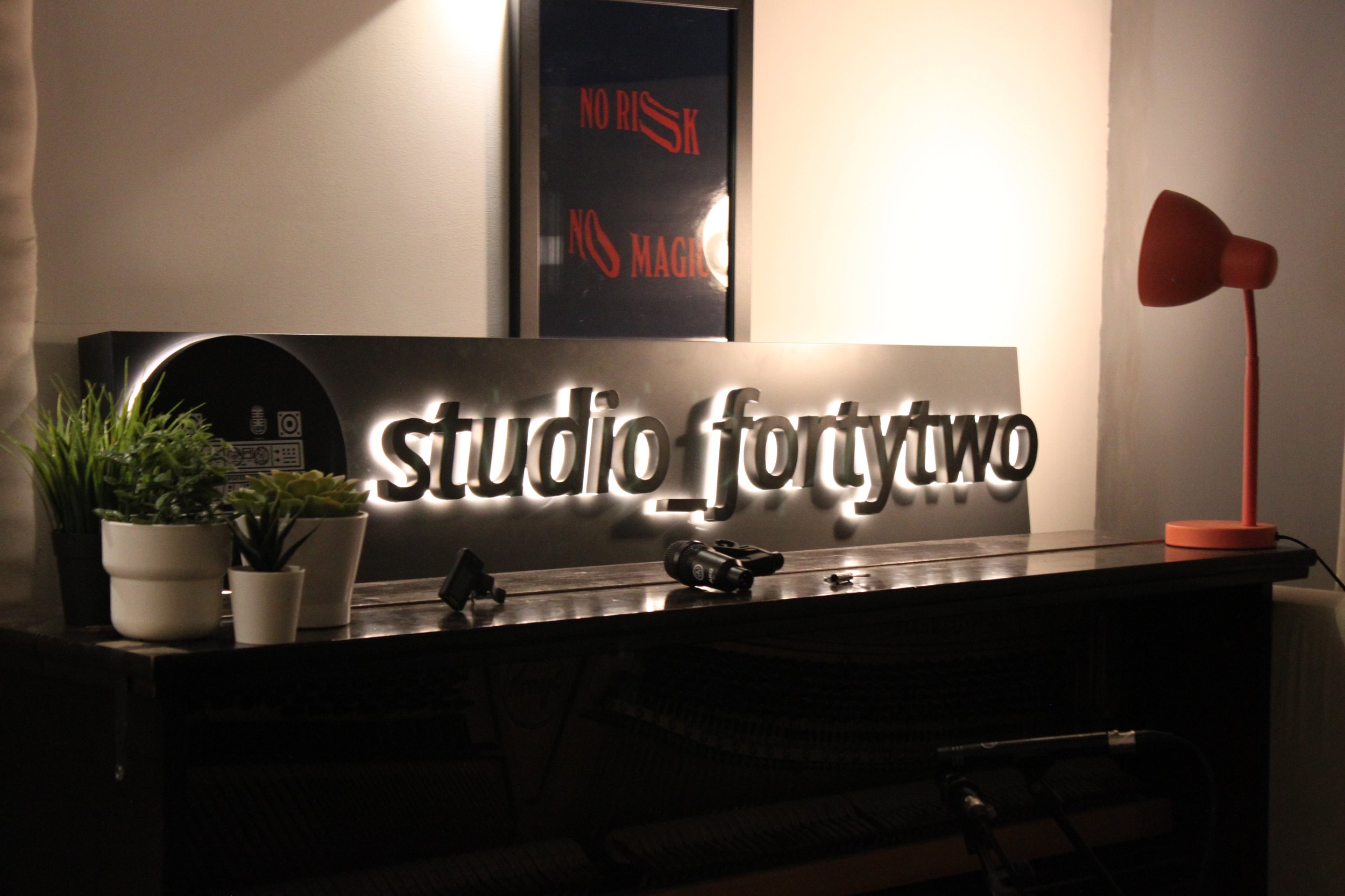 Interior view of a room with a backlit sign reading 'studio fortytwo' on a table, potted plants, a tripod, a flashlight, and a red lamp with a unique shade. A framed poster with red text says 'NO RISK NO MAGIC' on the wall.