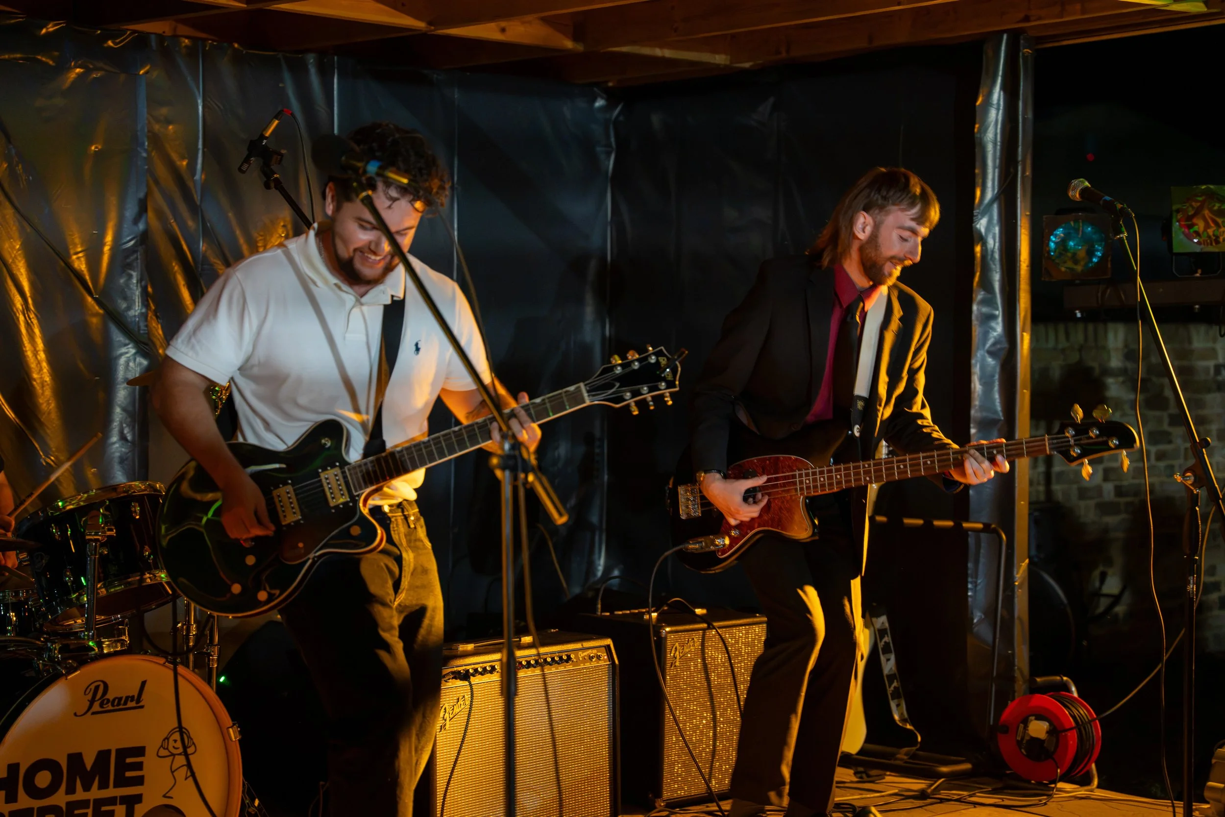 Two male musicians playing guitars on a stage, one with curly hair and a white shirt, the other with straight hair and a black jacket, illuminated by warm stage lighting. A drum set and amplifiers are visible in the background.