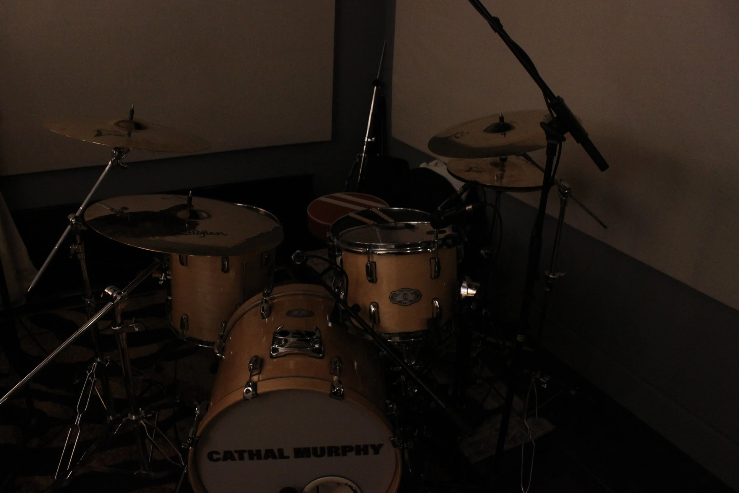 A drum set in a dimly lit room with a bass drum labeled 'Cathal Murphy,' and cymbals mounted on stands.