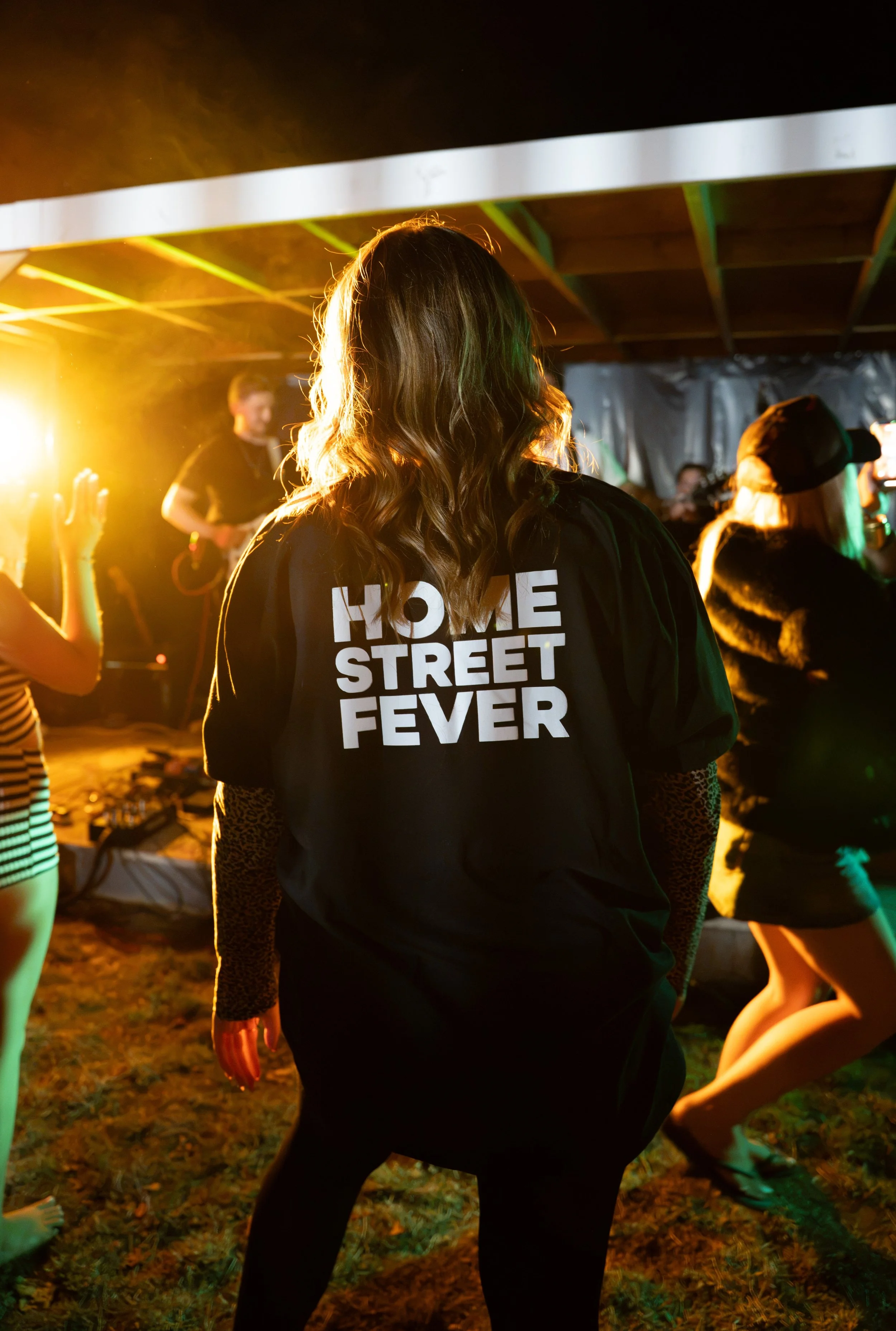 A woman with long wavy hair wearing a black shirt with white text that reads 'HOME STREET FEVER' is dancing at a dimly lit party or festival with other people in the background.