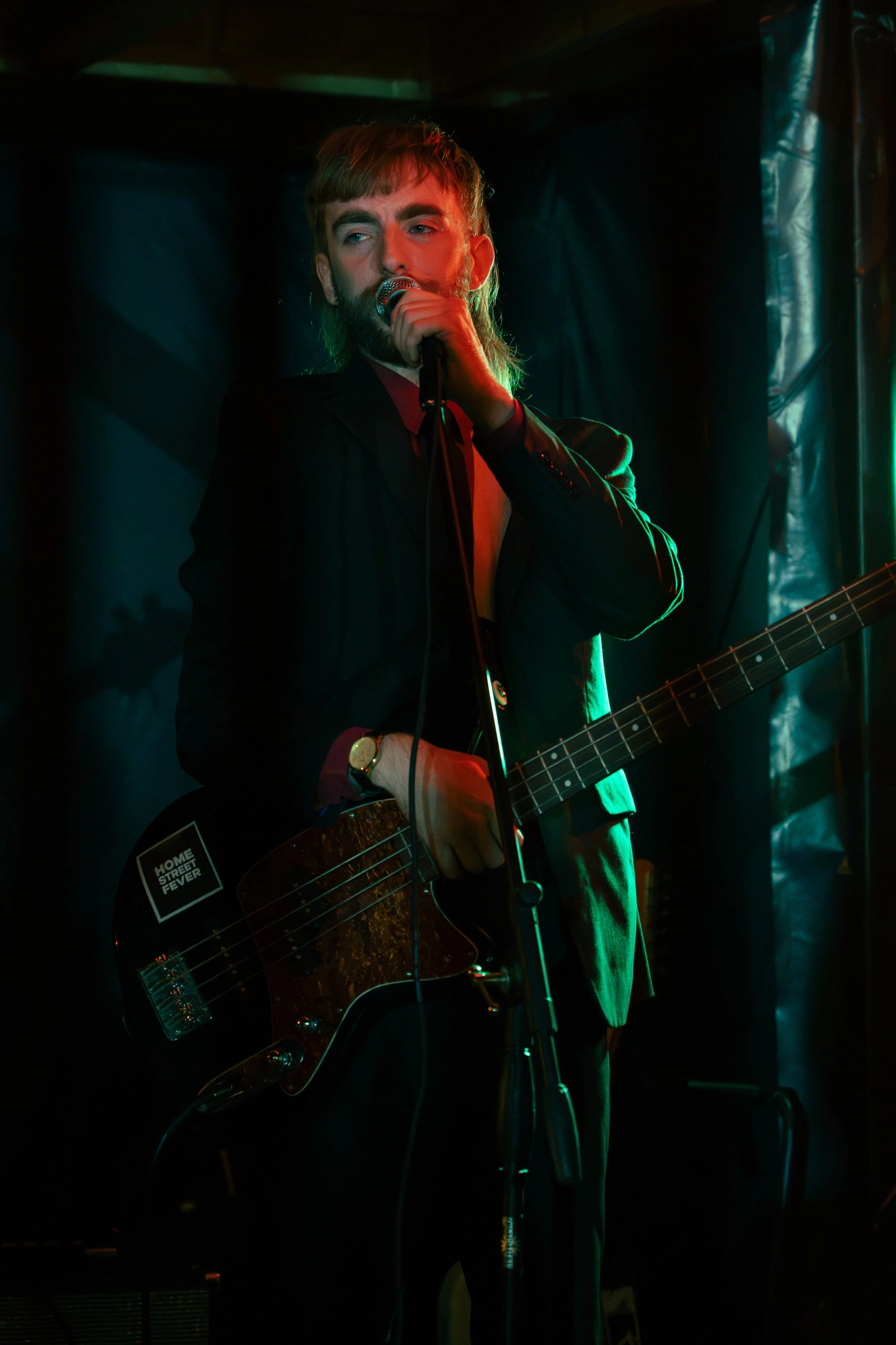 A man with a beard and brown hair singing into a microphone while playing an electric guitar on a dimly lit stage.