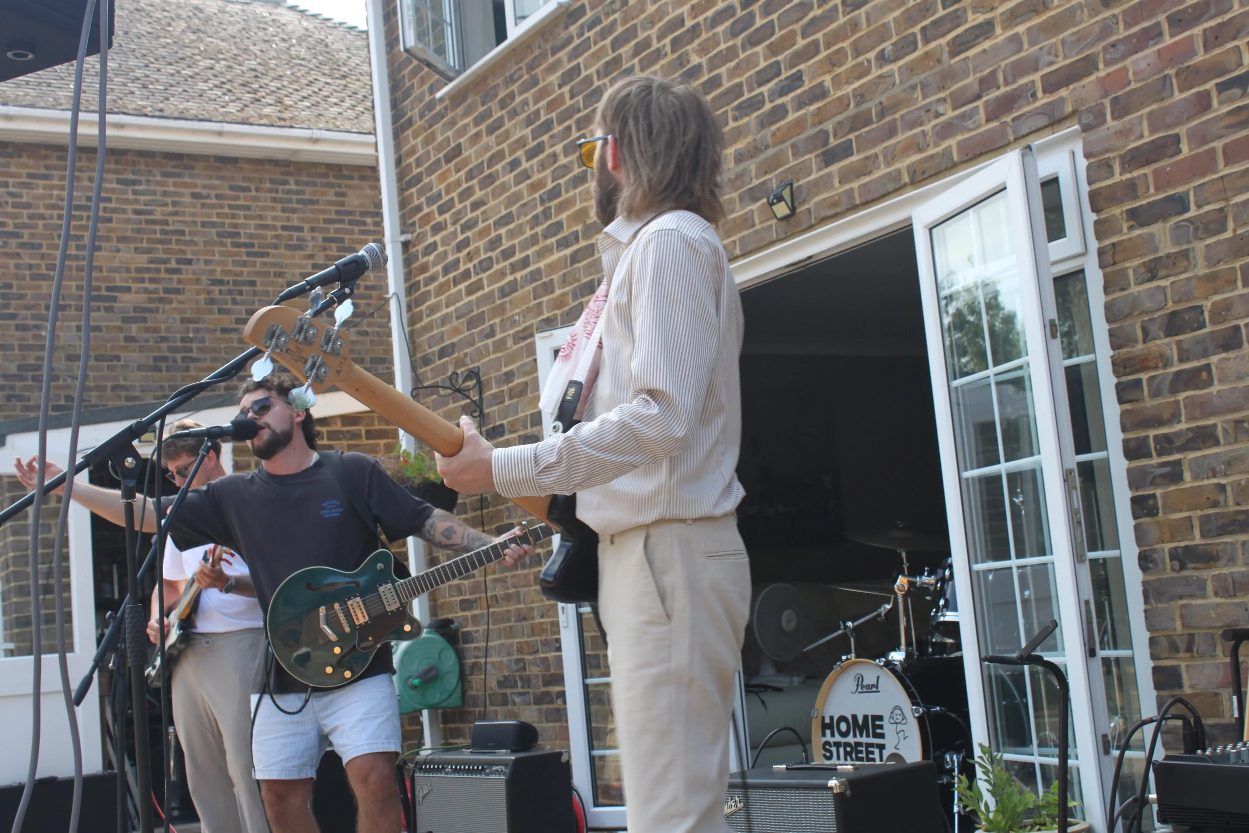 Musicians performing outdoors on a brick patio during daytime, with guitars and microphones, in front of a brick house with open windows and a drum set labeled "Home Street".