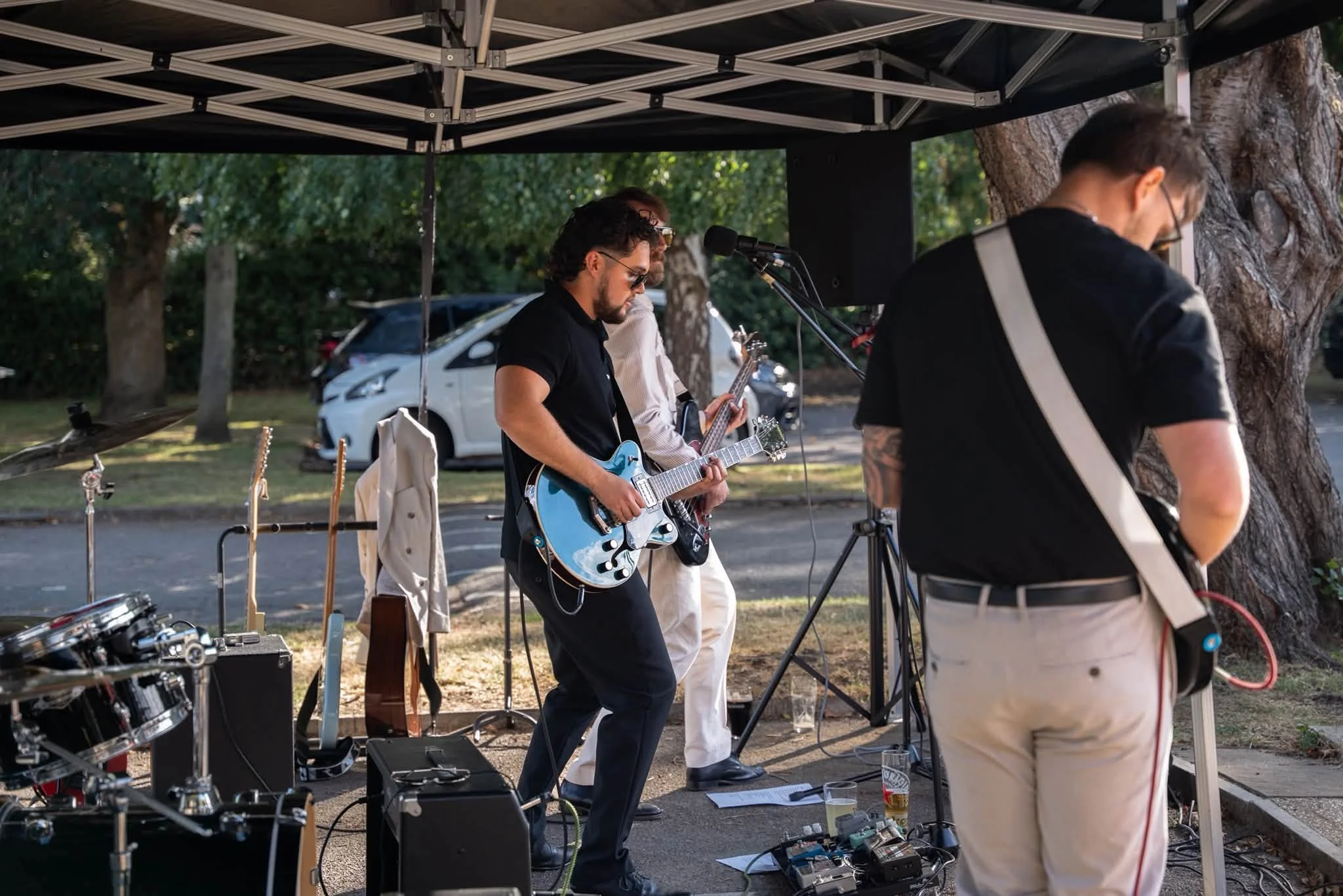 A group of musicians performing outdoors under a canopy, with trees and cars in the background. One person is playing an electric guitar, and others are preparing instruments.