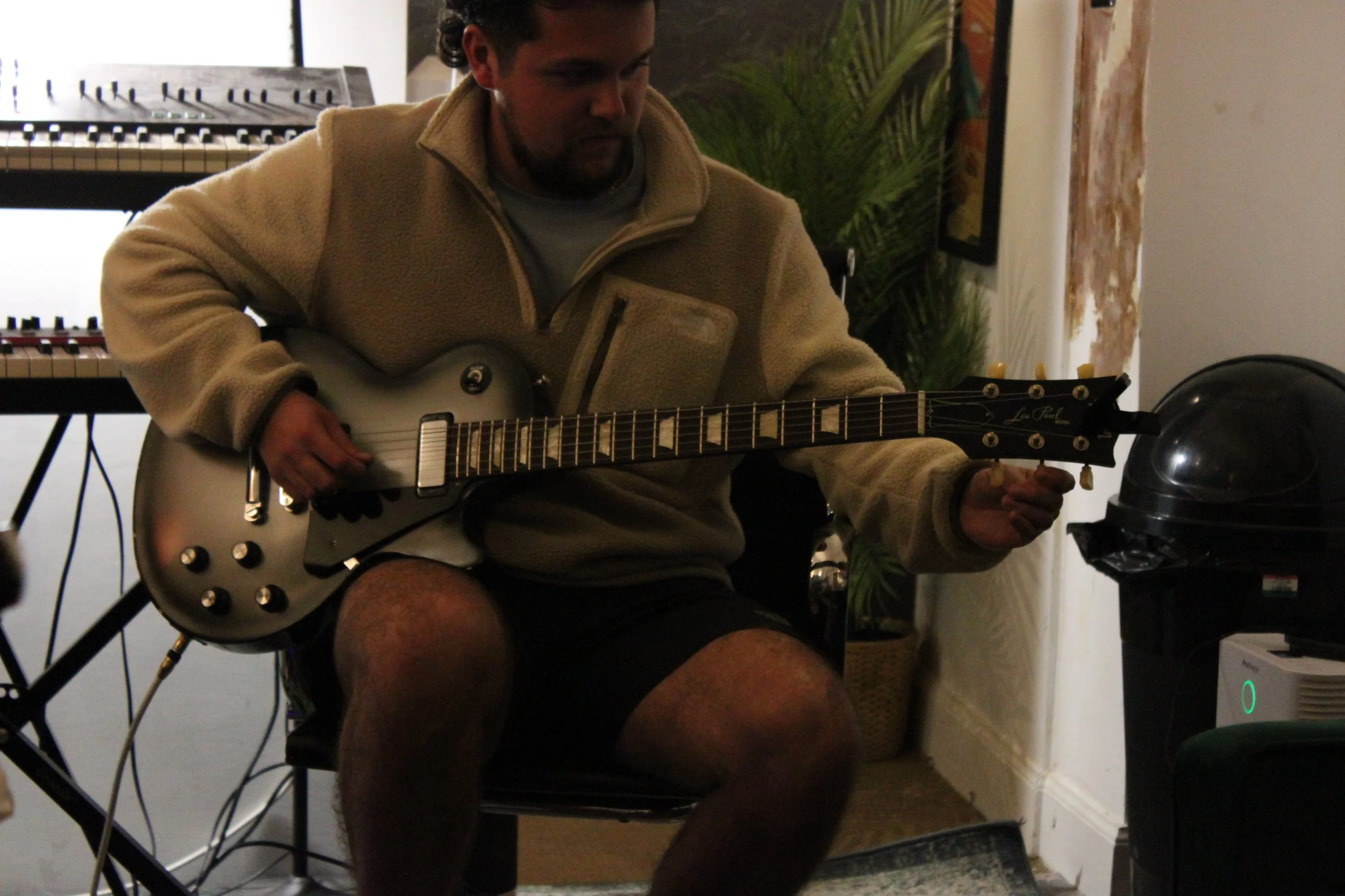 A man sitting in a room playing an electric guitar with a keyboard behind him and electronic equipment around.