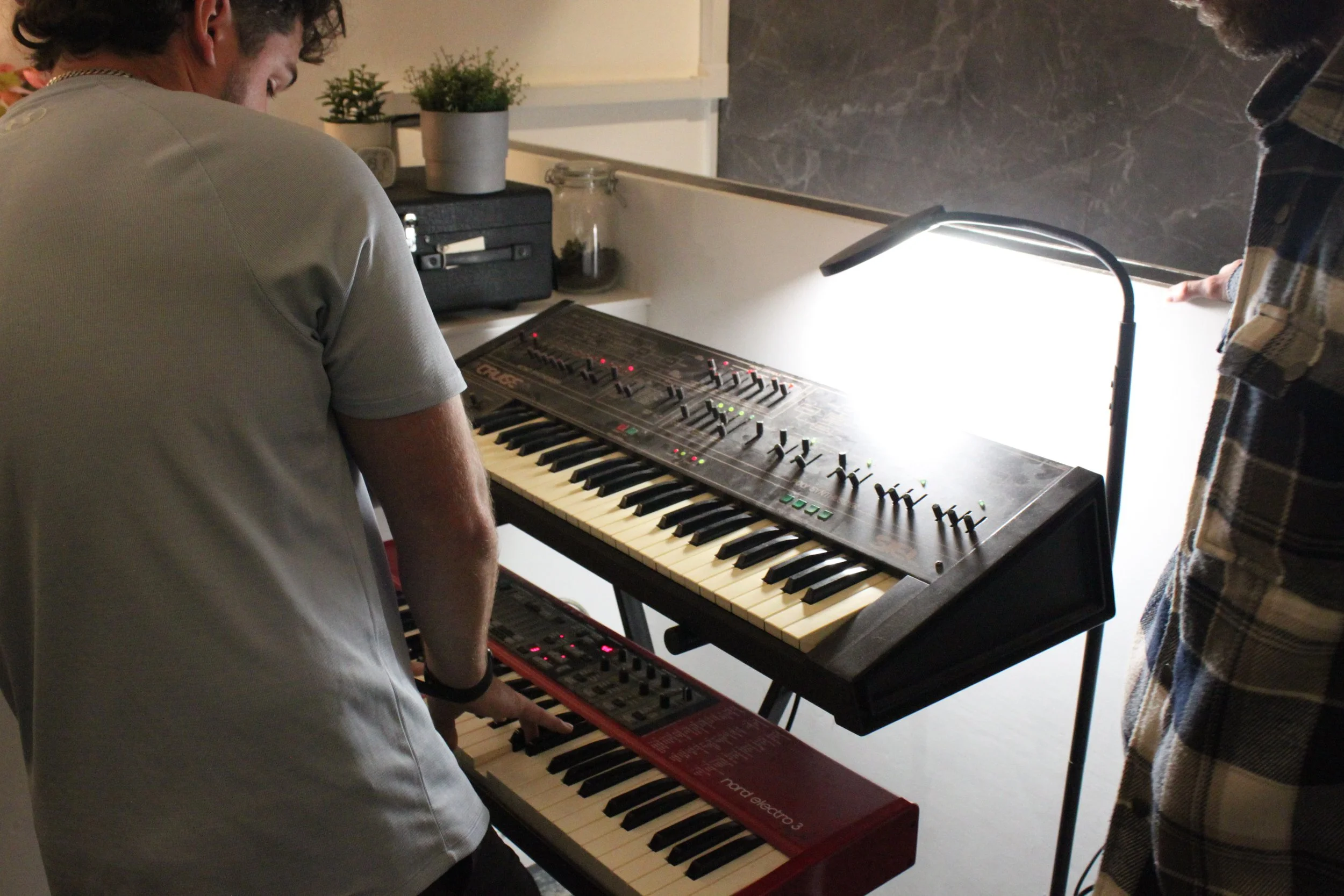 Two people are standing in front of an electronic keyboard with a digital control panel. One person, wearing a gray shirt, is playing the keyboard, while the other Person, dressed in a checkered shirt, observes. The scene appears to be in a room with