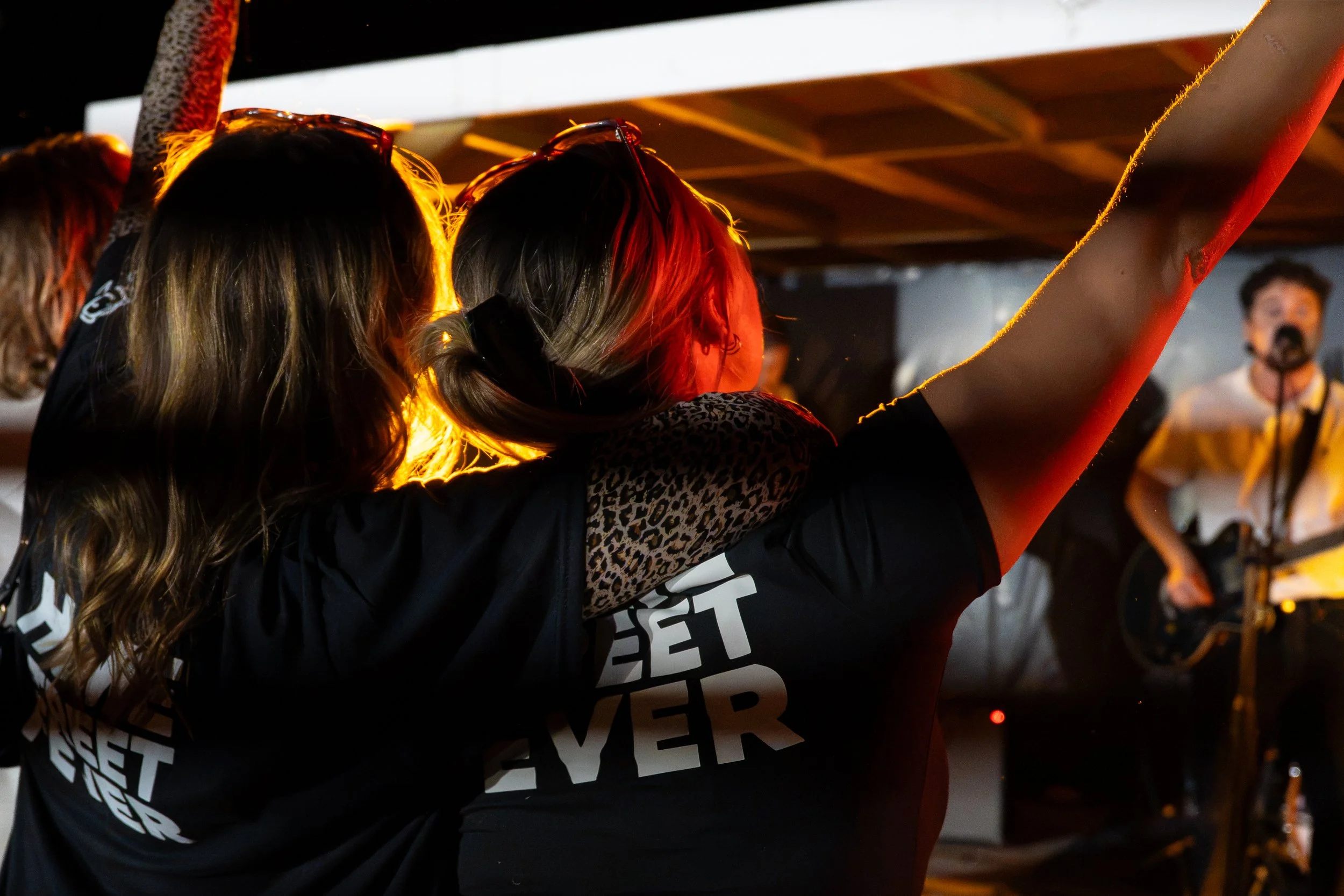 People at an event with one person raising a fist in the air, wearing sunglasses, under a structure with bright lighting.