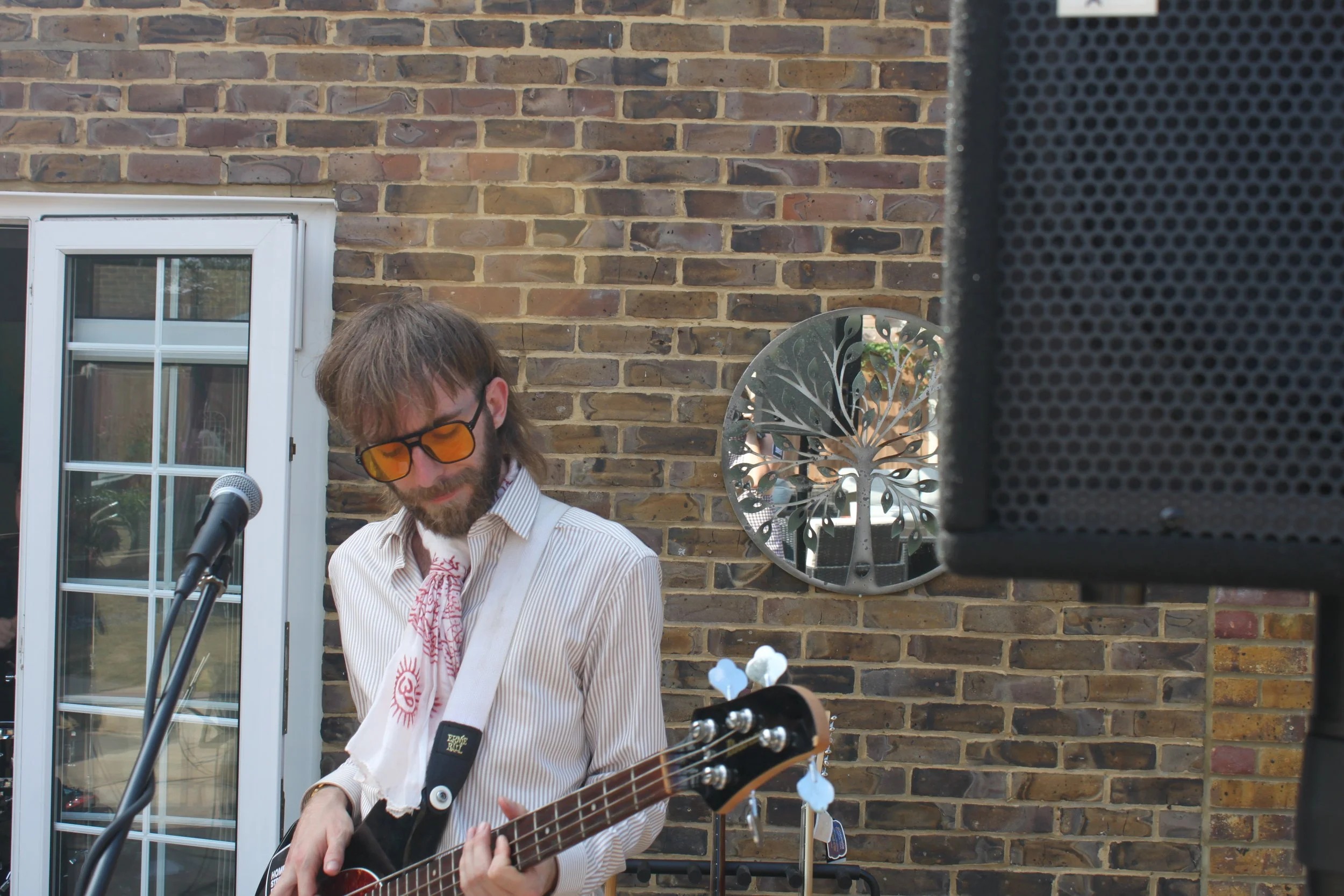 A man with a beard wearing sunglasses and a striped shirt playing an acoustic guitar outdoors. He is standing in front of a brick wall window with a decorative mirror and tree-shaped wall art. Part of a black speaker is visible in the foreground.