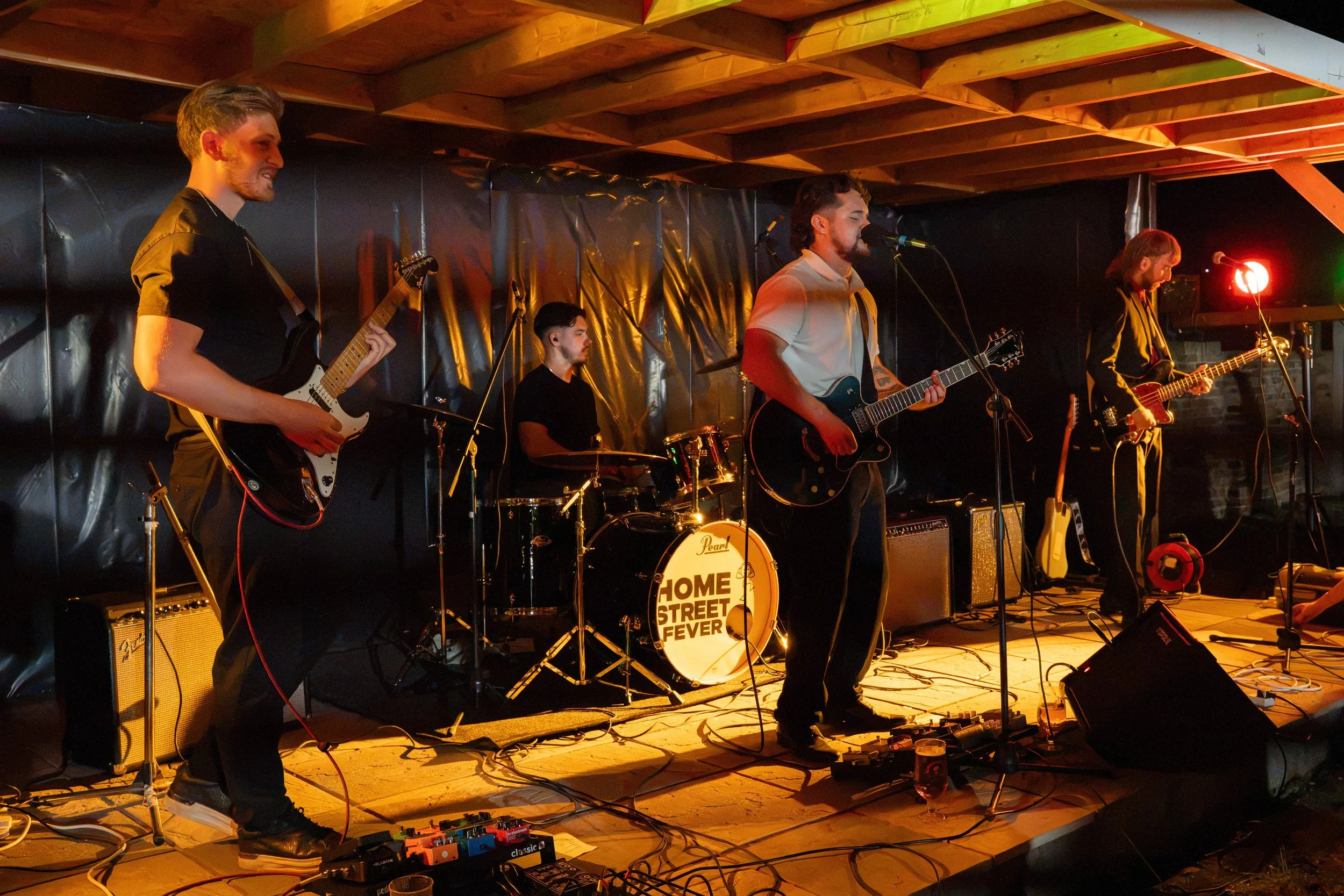 A band of five musicians performing on a stage in a dimly lit setting, playing guitars, drums, and singing into microphones, with a drum set displaying a sign that reads 'Home Street Fever'.