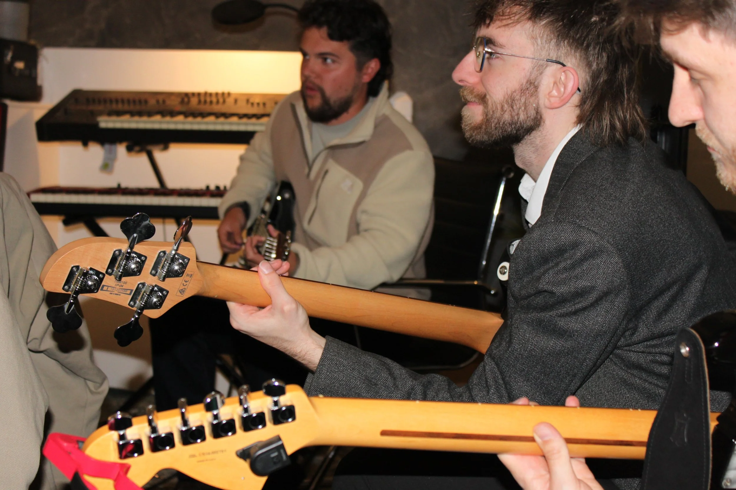Three men sitting together in a music studio, playing guitars. One man is wearing glasses and a gray blazer, smiling. The second man has dark hair and a beard, wearing a cream colored jacket, playing an electric guitar. The third person is partially 
