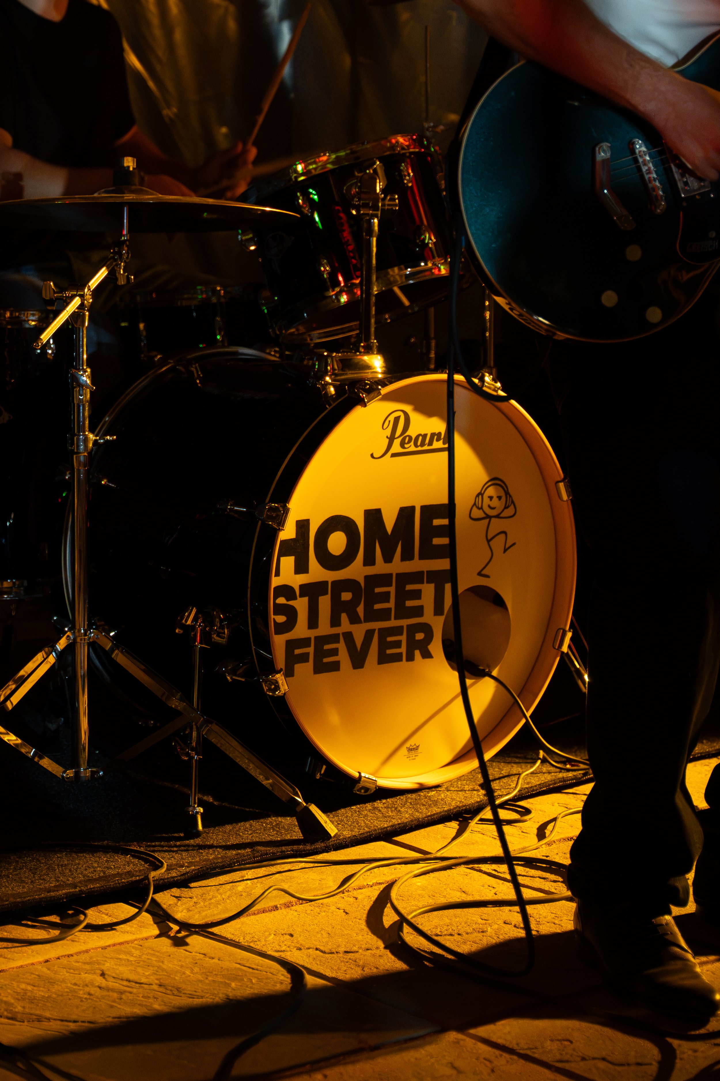 Close-up of a drum set on stage, with a bass drum displaying the words "Home Street Fever" and a small cartoon figure. A musician's arm is visible, playing the guitar. Cables are scattered on the stage floor.