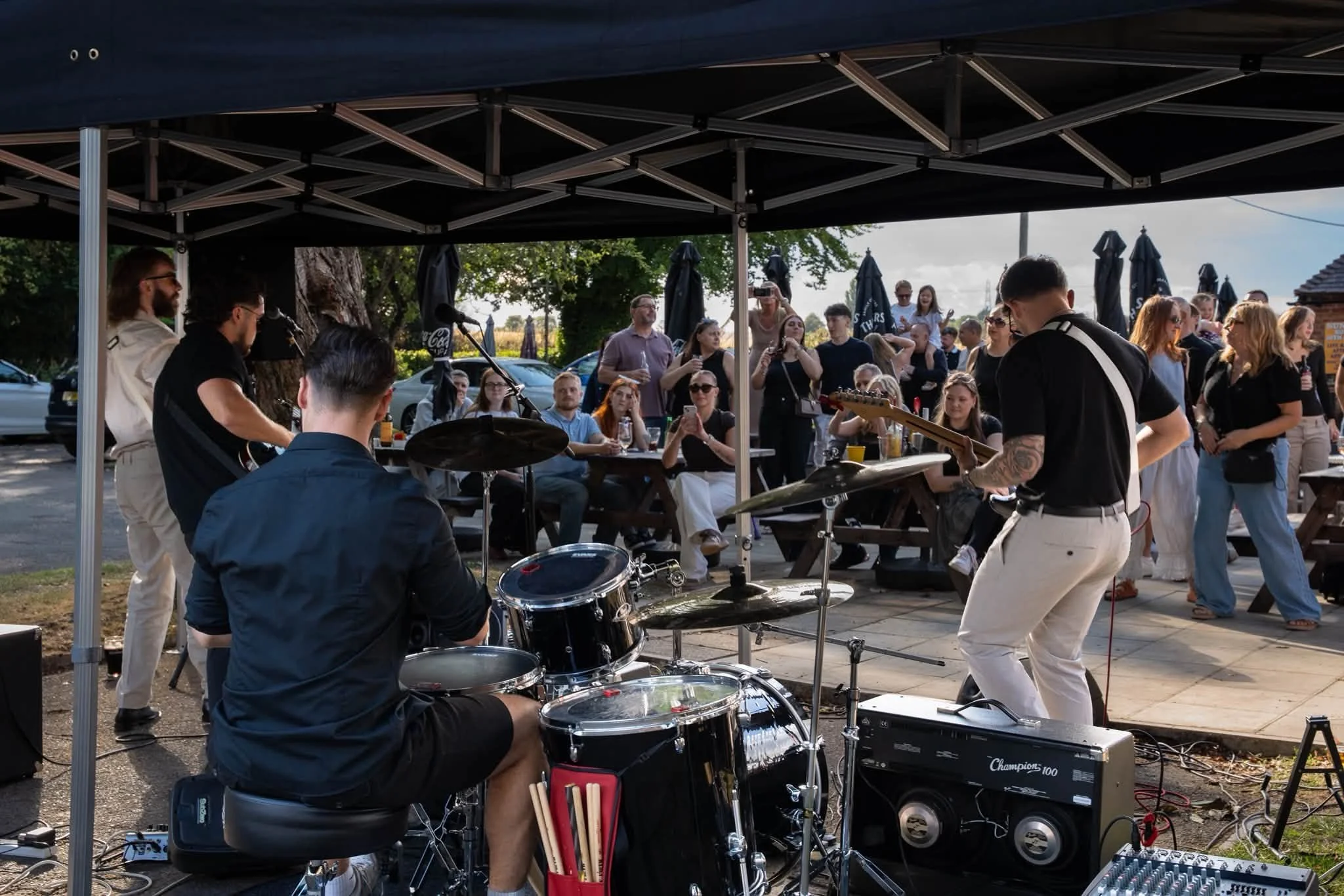 A live outdoor music performance with a band under a black canopy, surrounded by an audience, some taking photos or videos. The band includes a drummer, a guitarist, and a singer, with people enjoying the event in a sunny setting.