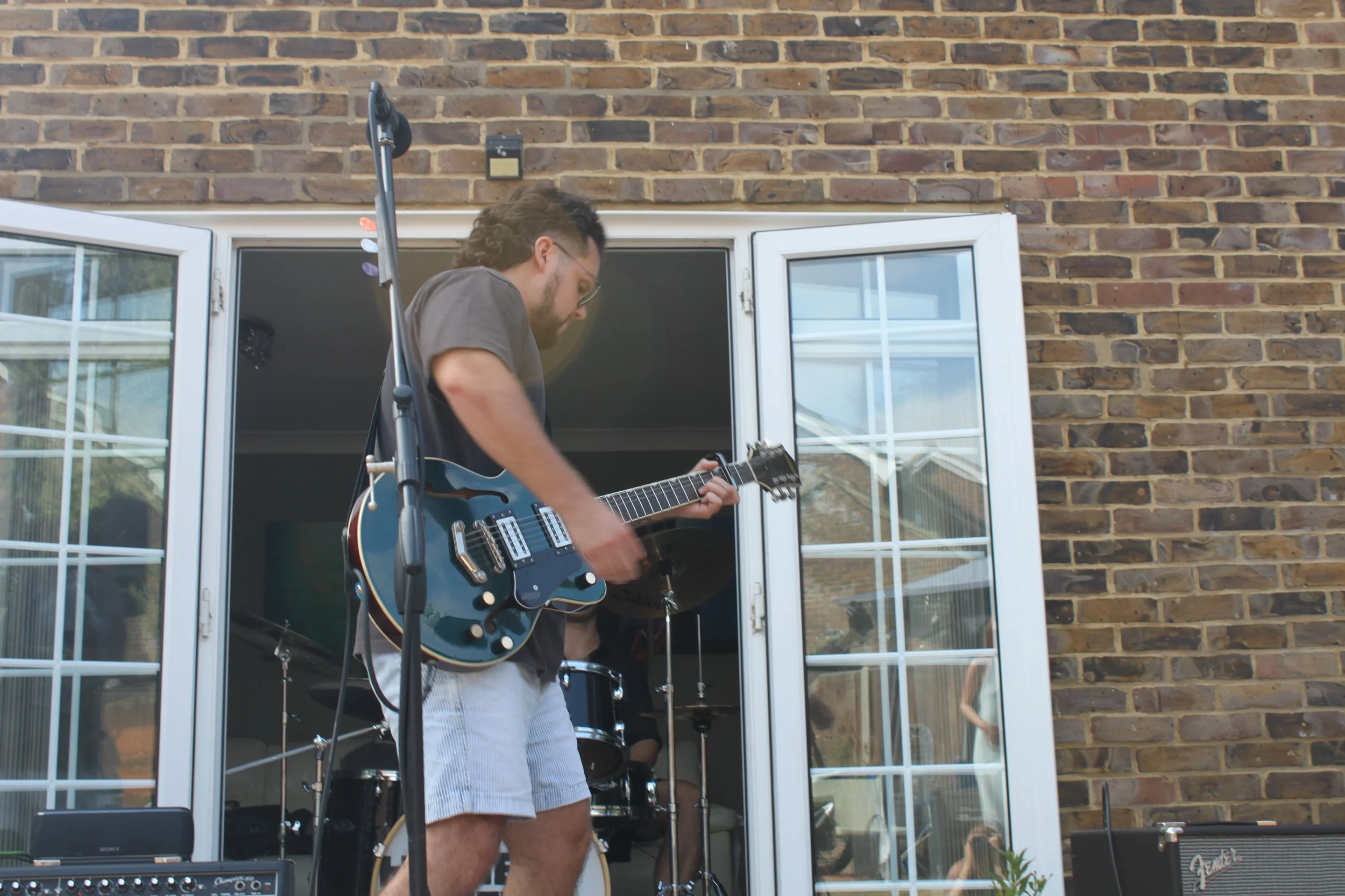 A man with glasses playing an electric guitar during an outdoor performance on a patio with a brick wall. There is musical equipment around him, including a black amplifier, a drum set, and a guitar stand. The scene is near a sliding glass door, refl