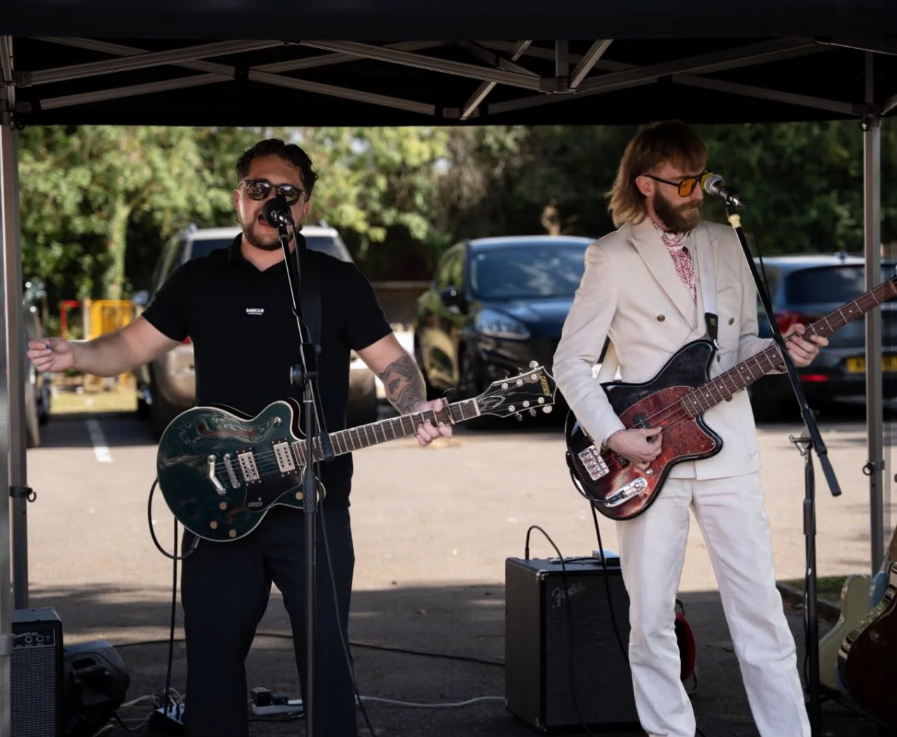 Two men perform as a band under a canopy, each playing an electric guitar and singing into microphones, with parked cars and trees in the background.