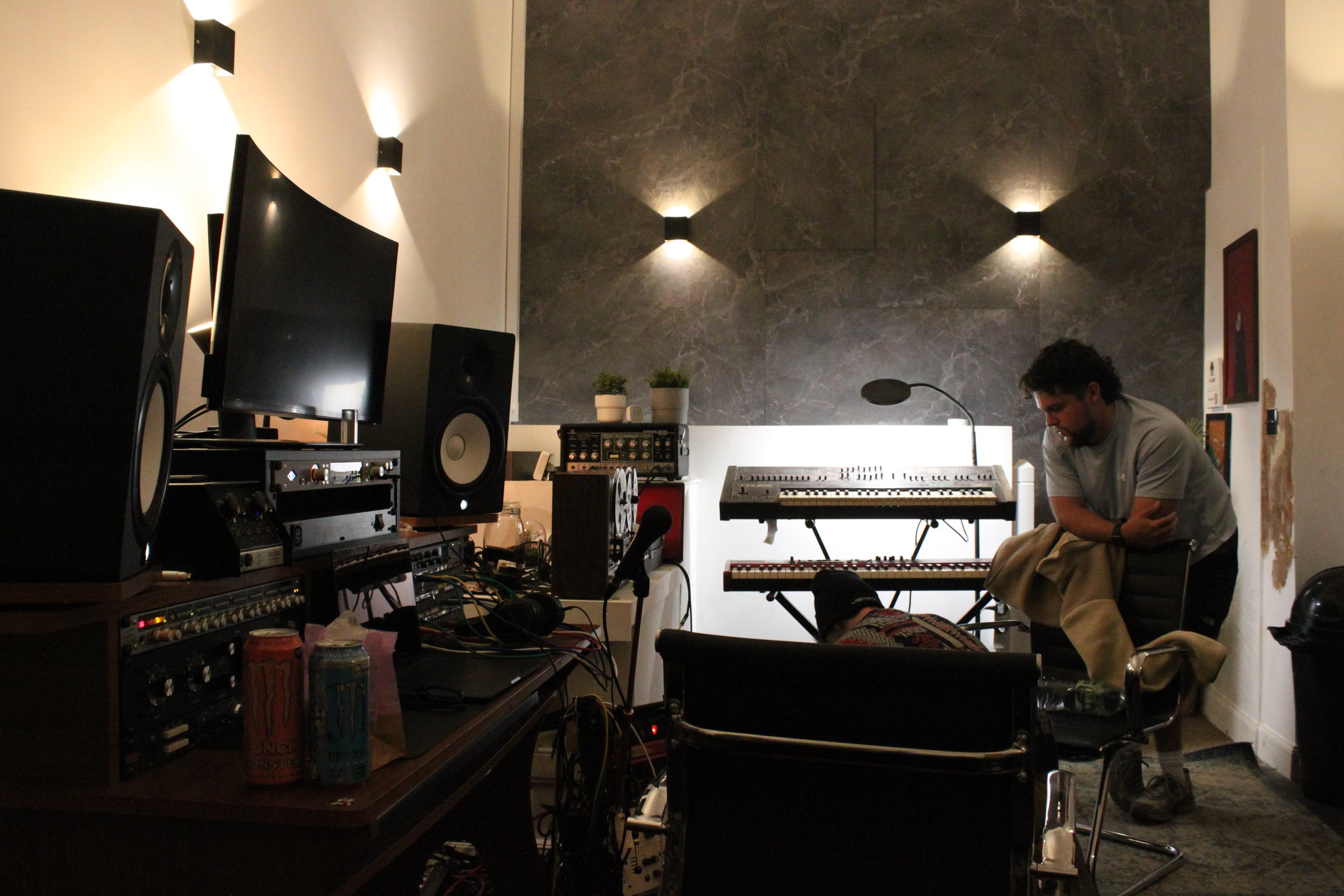 A music studio with various equipment including speakers, monitors, and synthesizers. A man is sitting with bent downward head in front of a keyboard, next to a person in a wheelchair.