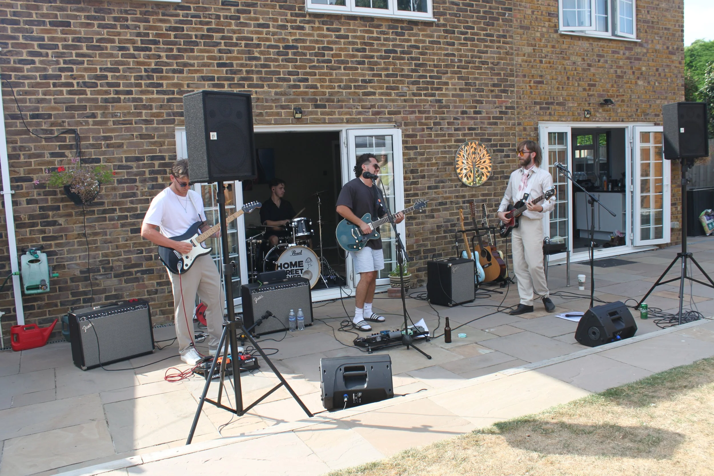 A band performing outside in a backyard. Four musicians are playing guitars and drums, with speakers and microphones set up. The house has brick walls and open French doors.