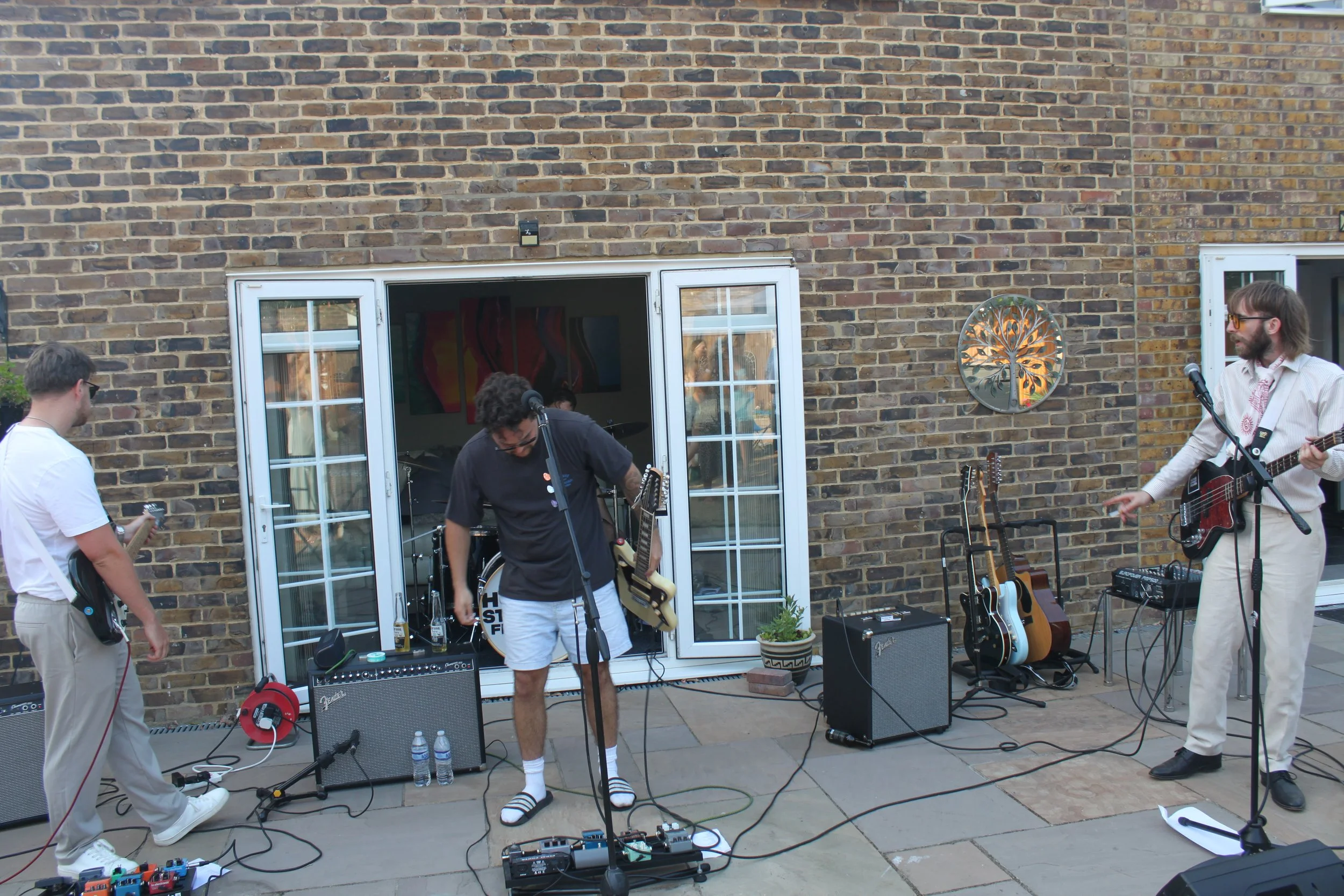 A group of musicians performing outside in a backyard with a brick wall background. They have guitar and music equipment, and one person is singing into a microphone while playing the guitar.