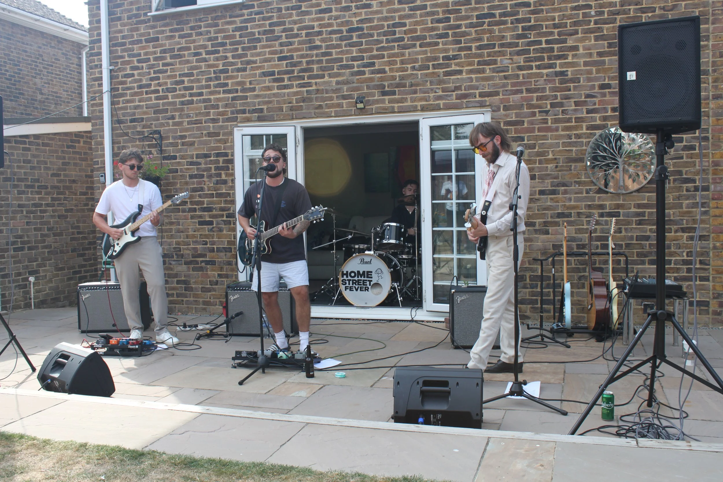 Four young men are performing a live music set outdoors in front of a brick wall. They are playing electric guitars and a drum set, with microphones and amplifiers set up around them. One man is singing into a microphone, while the others are focused