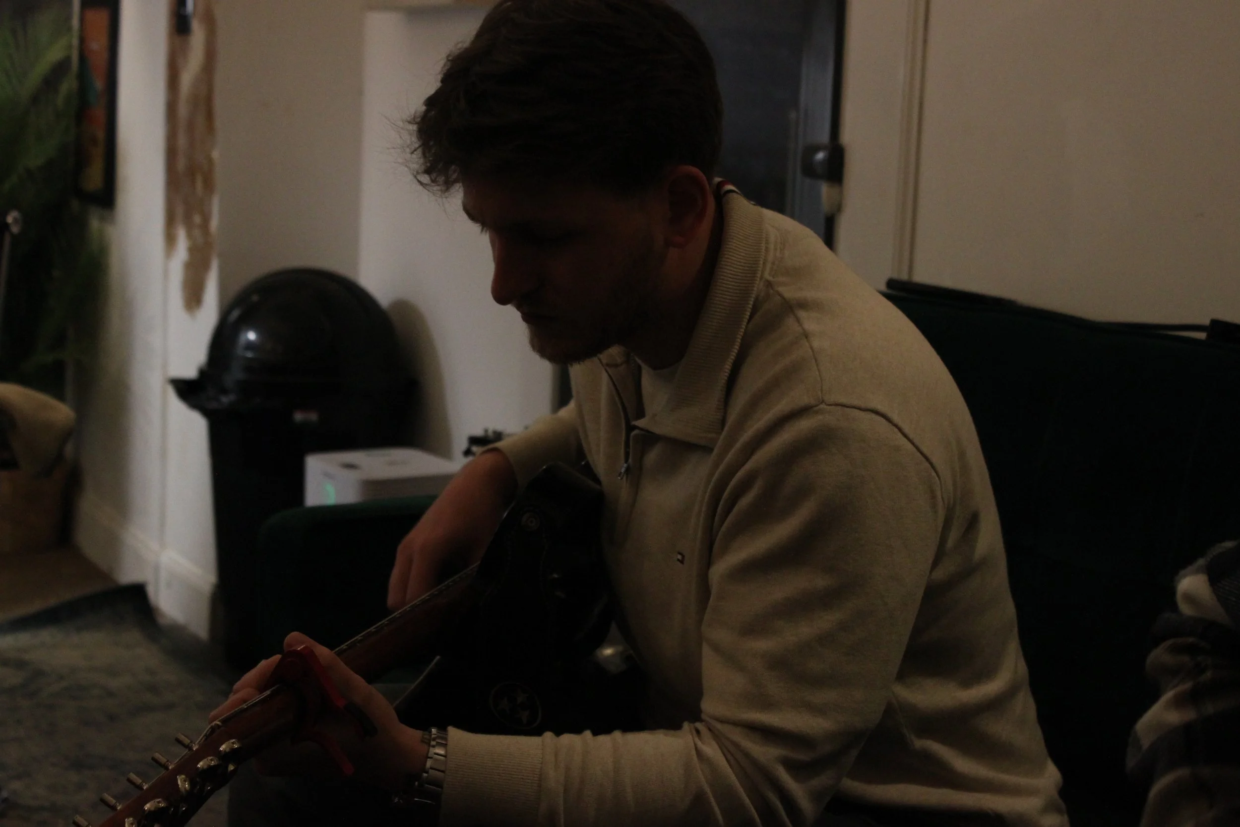 A man with dark hair, a beard, and a light-colored jacket is sitting on the floor playing an acoustic guitar in a dimly lit room.