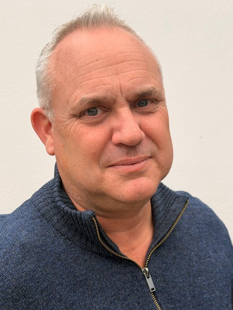 Close-up of a middle-aged man with short gray hair, wearing a dark blue zip-up sweater, standing against a plain light-colored wall.