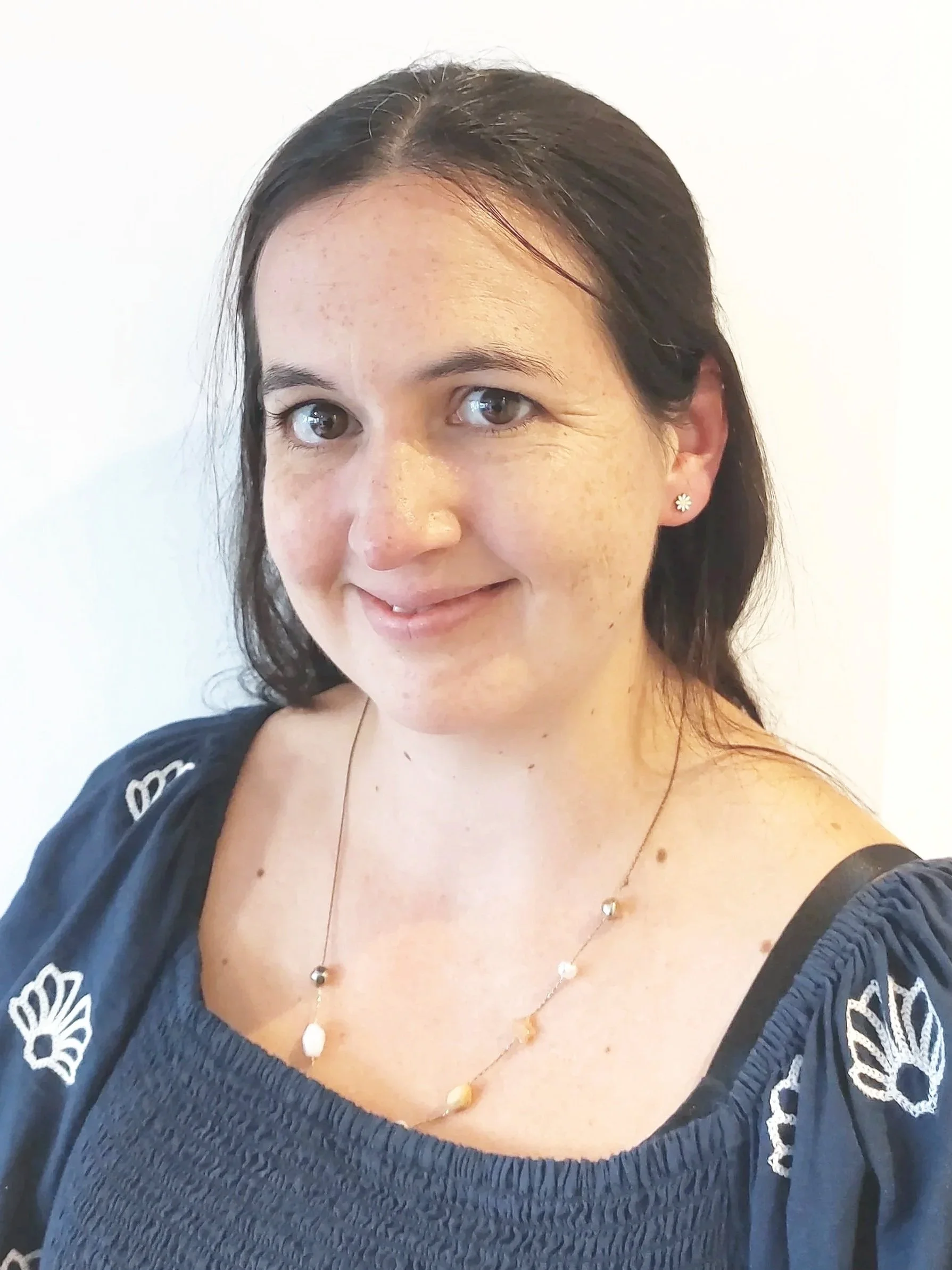 Close-up of a woman's face and upper body, wearing a black embroidered top, delicate necklace with white stones, and small flower-shaped earrings, against a plain white background.