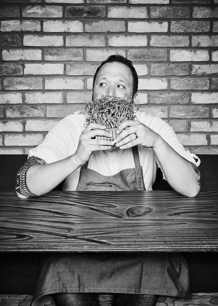 Man with a beard and tattoos sitting at a wooden table, holding a small potted plant with roots near his face, against a brick wall background, in black and white.