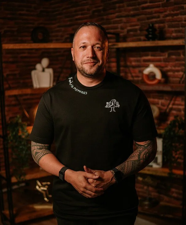 Man standing indoors in front of a brick wall with shelves, wearing a black t-shirt with a tree graphic and text, with tattoos on his arms.