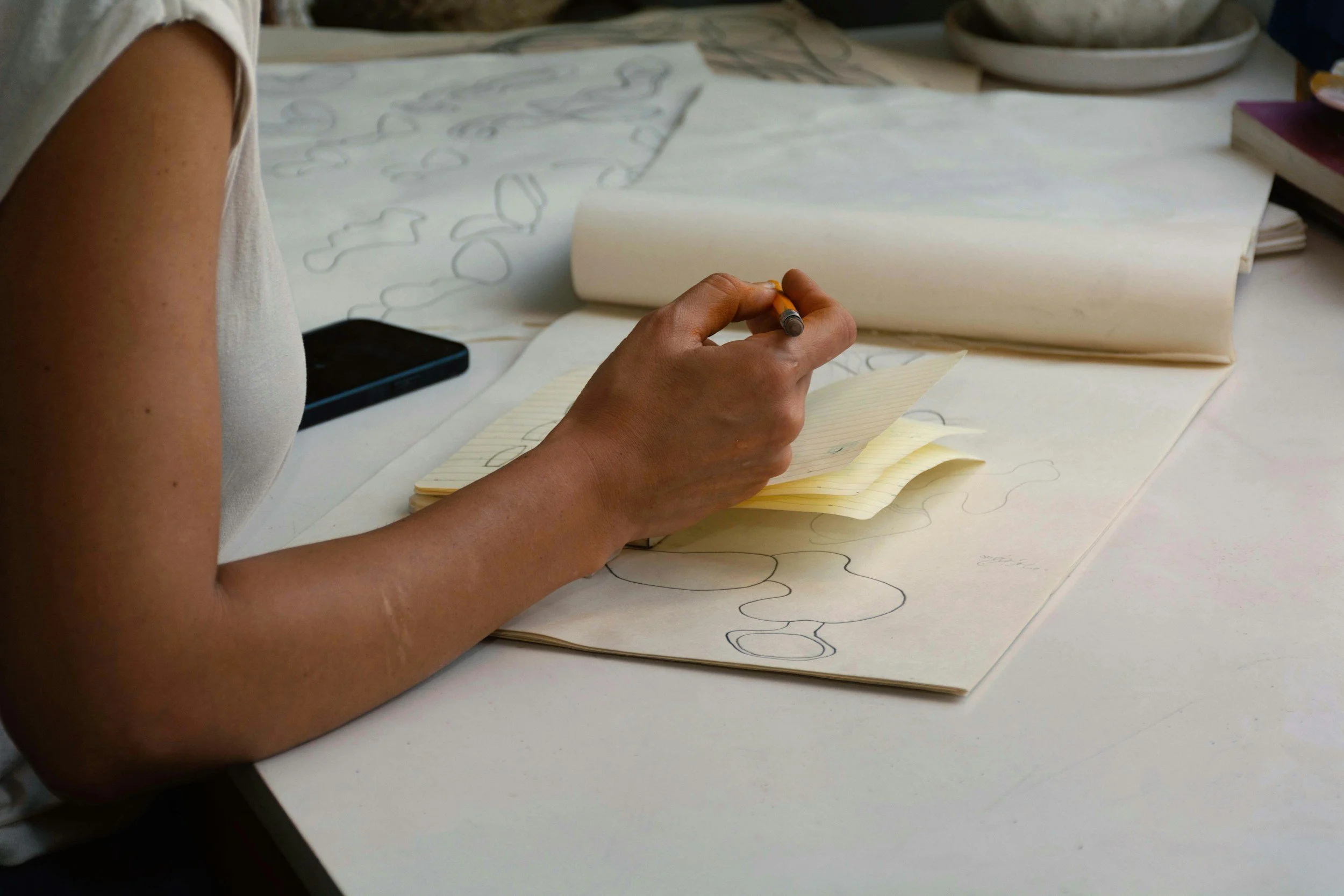 A person sketching abstract shapes on a large sheet of paper at a cluttered desk, with sticky notes, a smartphone, and a bowl of stones nearby.
