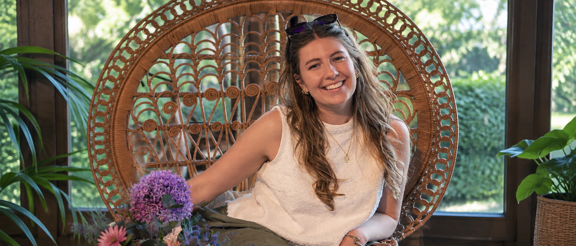 A smiling young woman with long, wavy hair, wearing a white sleeveless top, sitting in a large wicker peacock chair. She has sunglasses resting on her head and is surrounded by green potted plants. There are colorful flowers in front of her and a window with a view of greenery in the background.