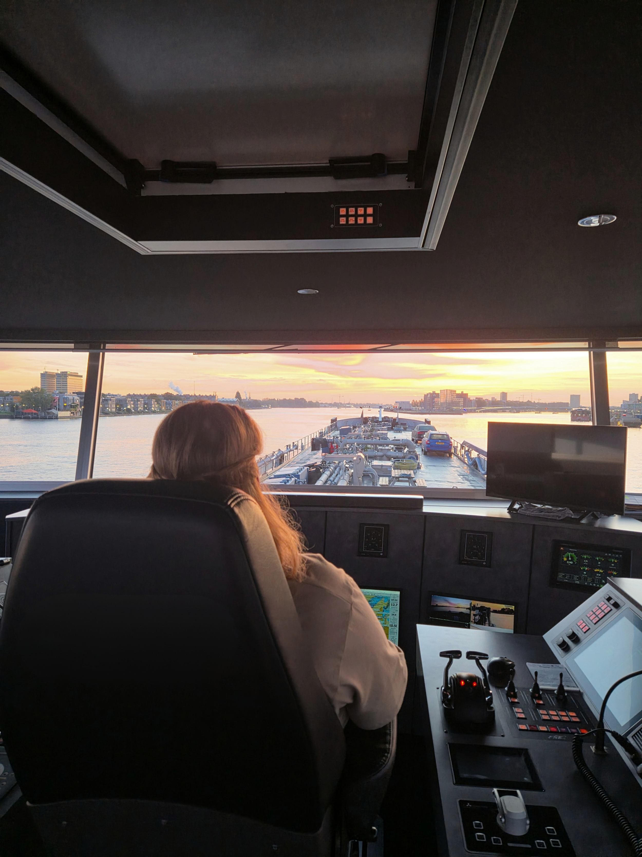 De afbeelding toont de cockpit van een schip met uitzicht op de rivier tijdens zonsondergang. Er is een persoon die aan de ene kant van het schip zit en de navigatieapparatuur en bedieningspanelen worden weergegeven. Vrouwen in de binnenvaart.